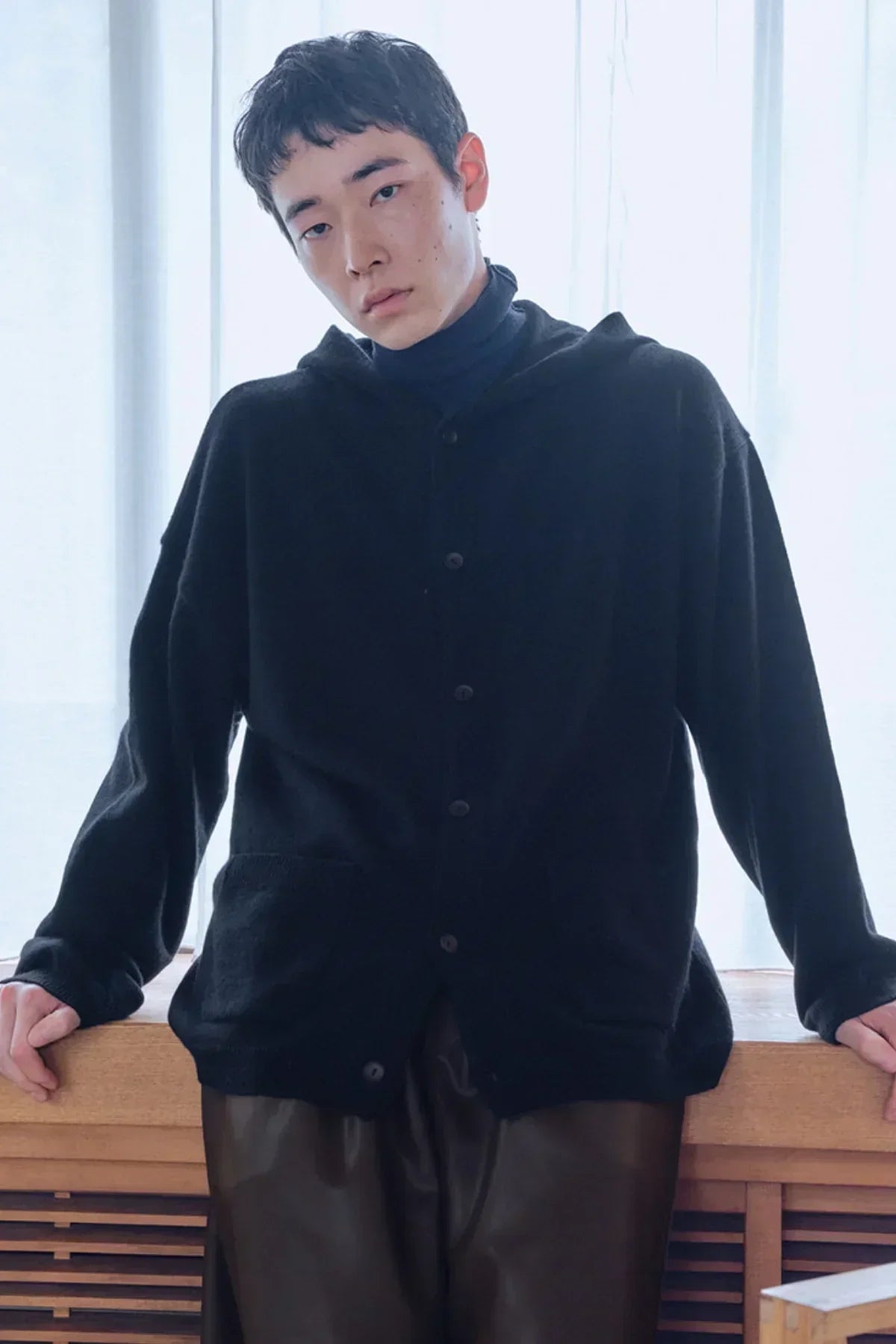 Young man wearing black knit hooded cardigan and dark pants indoors by wooden ledge