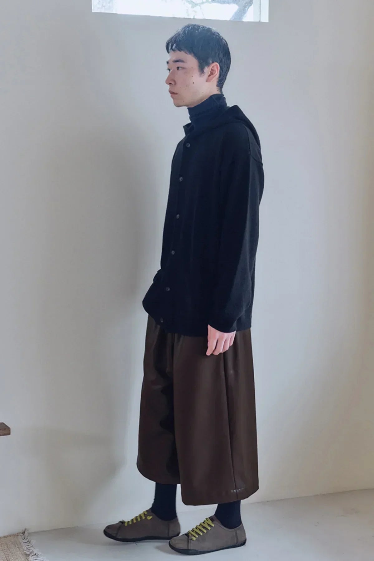 Side view of a model wearing black knit hooded cardigan, brown wide-leg pants, and gray sneakers indoors