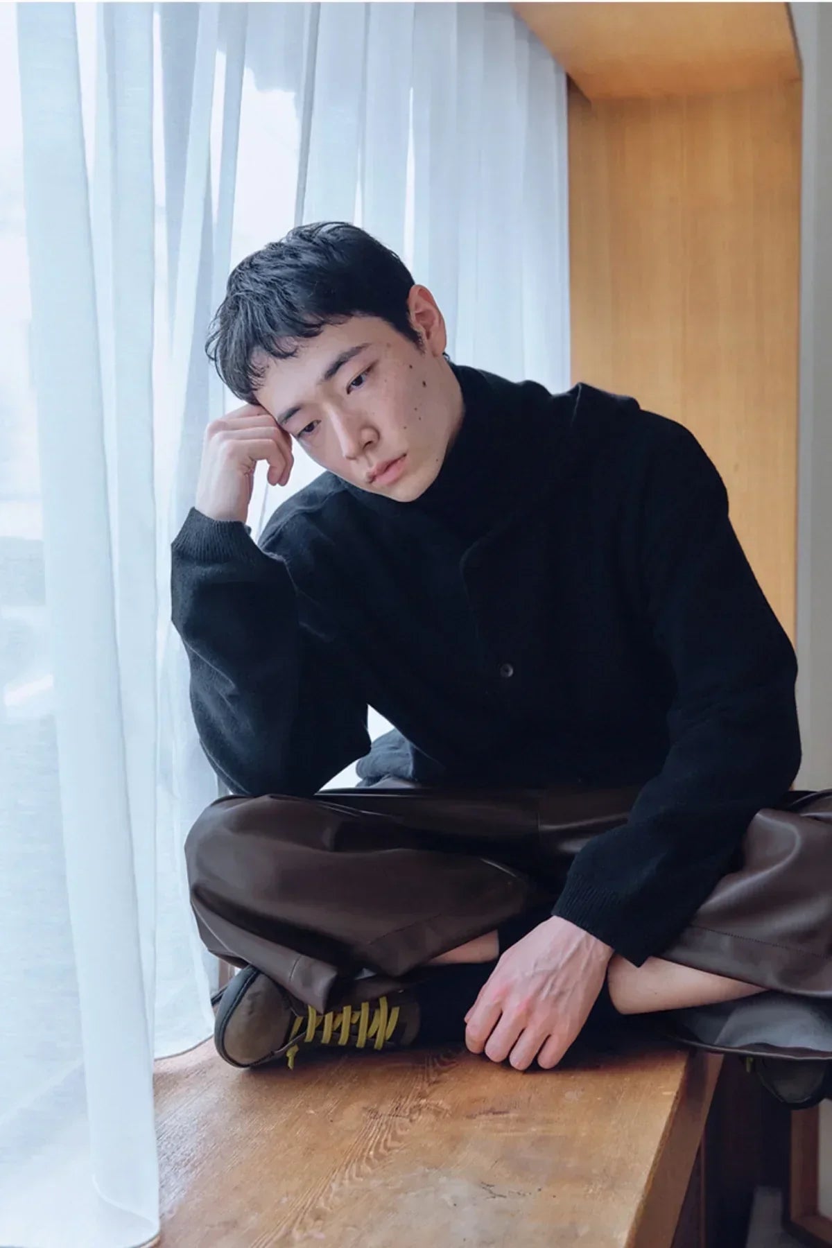 Young man in black knit sweater and brown pants sitting cross-legged on wooden windowsill with sheer white curtains