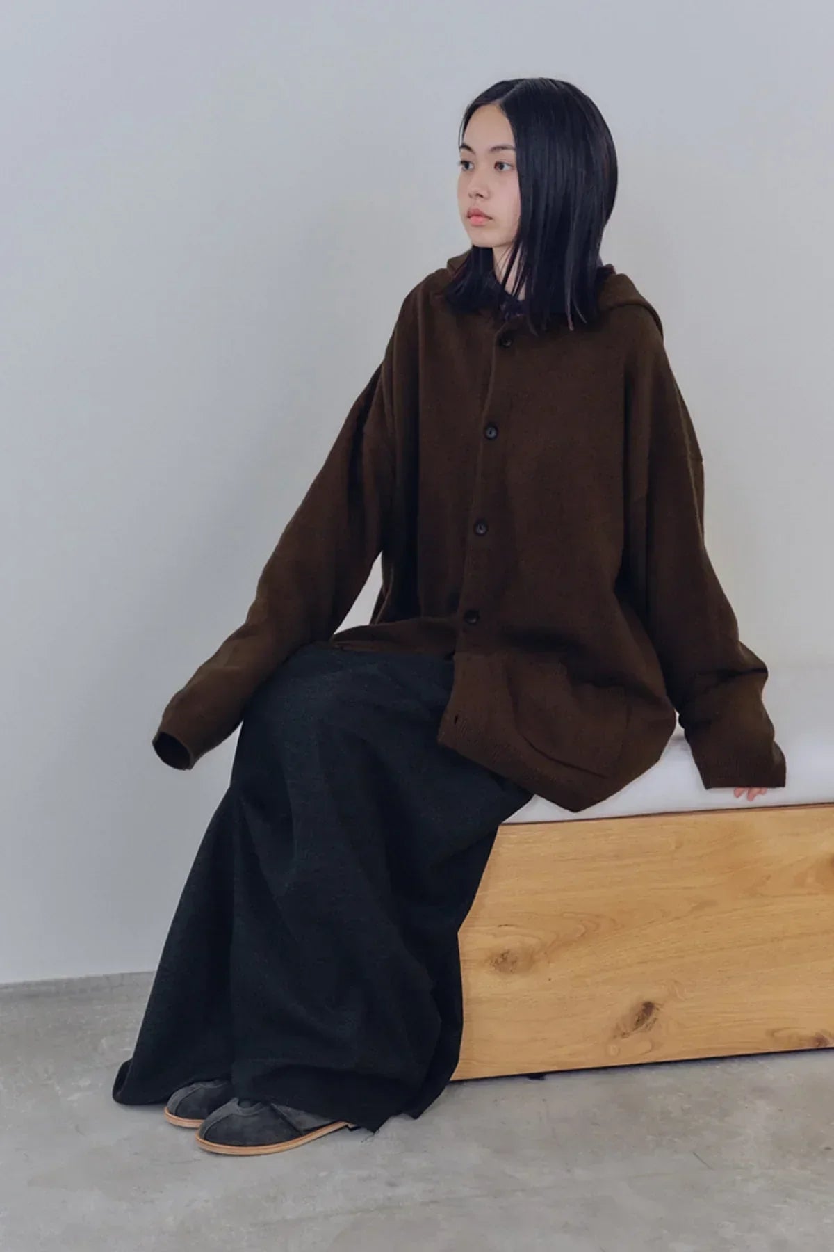 Woman wearing oversized brown knit hooded cardigan and dark wide-leg pants sitting on wooden bench