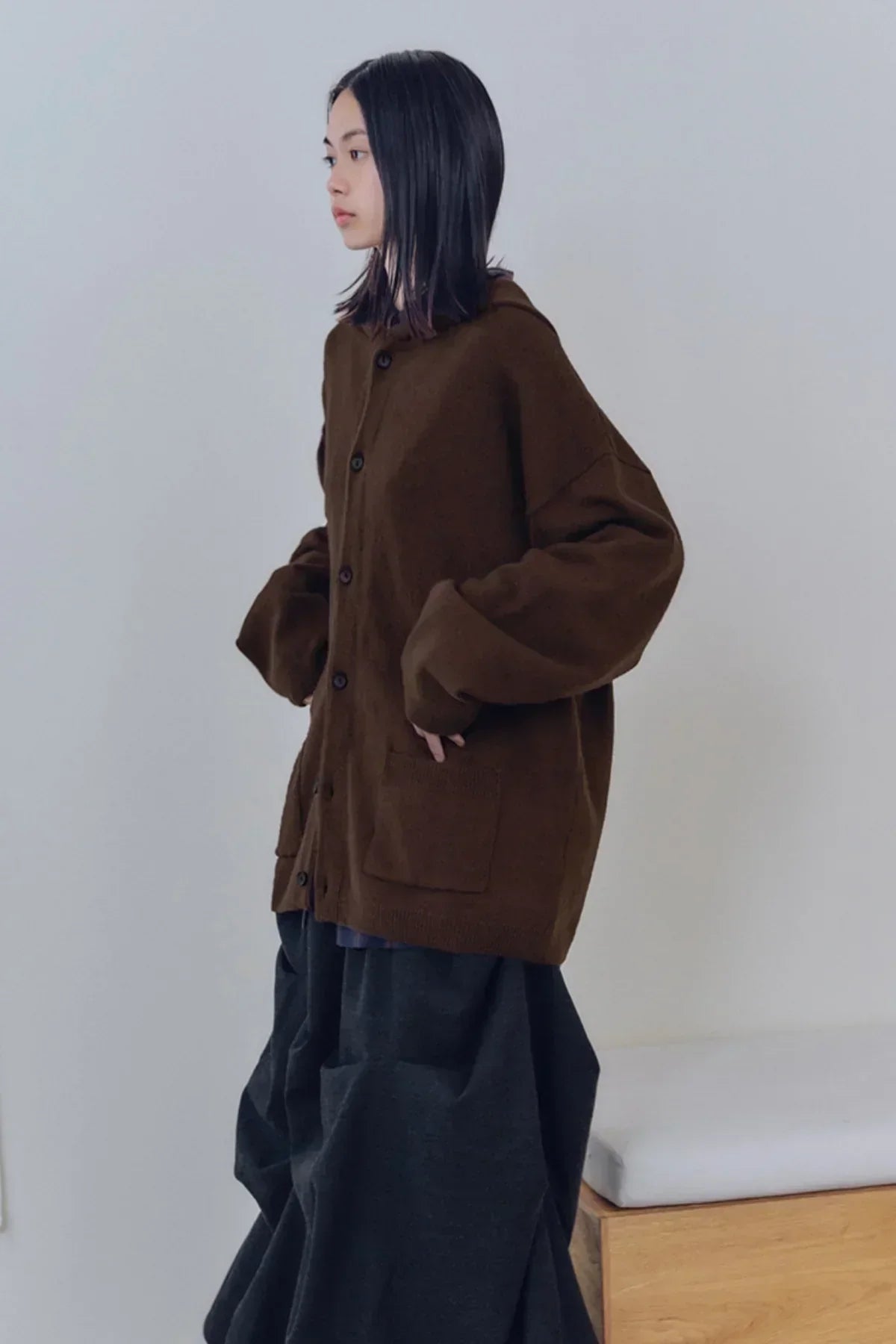 Side view of woman wearing brown knit buttoned cardigan with pockets and loose dark pants indoors