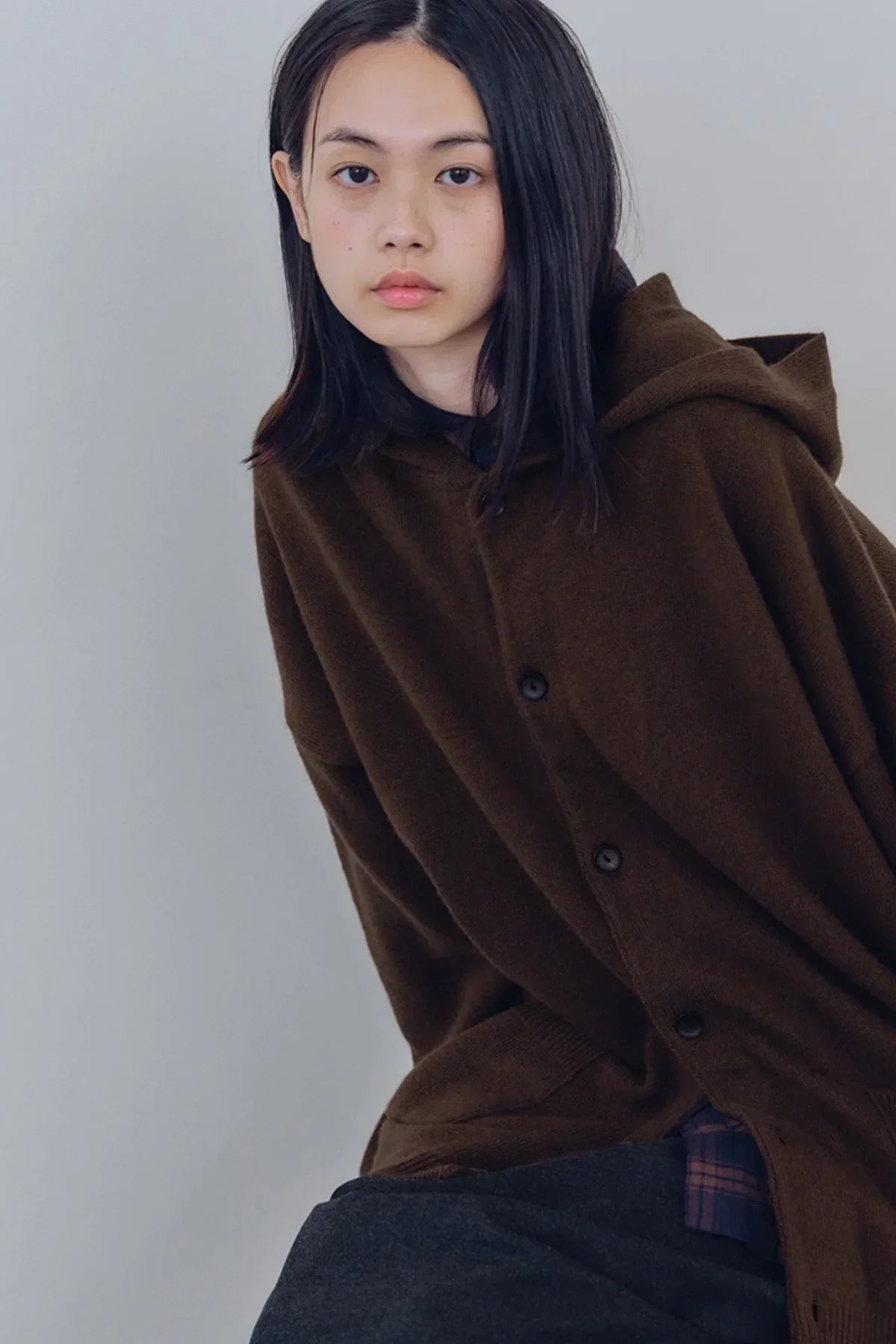 Young person wearing a brown knit hooded cardigan with dark pants against a plain backdrop