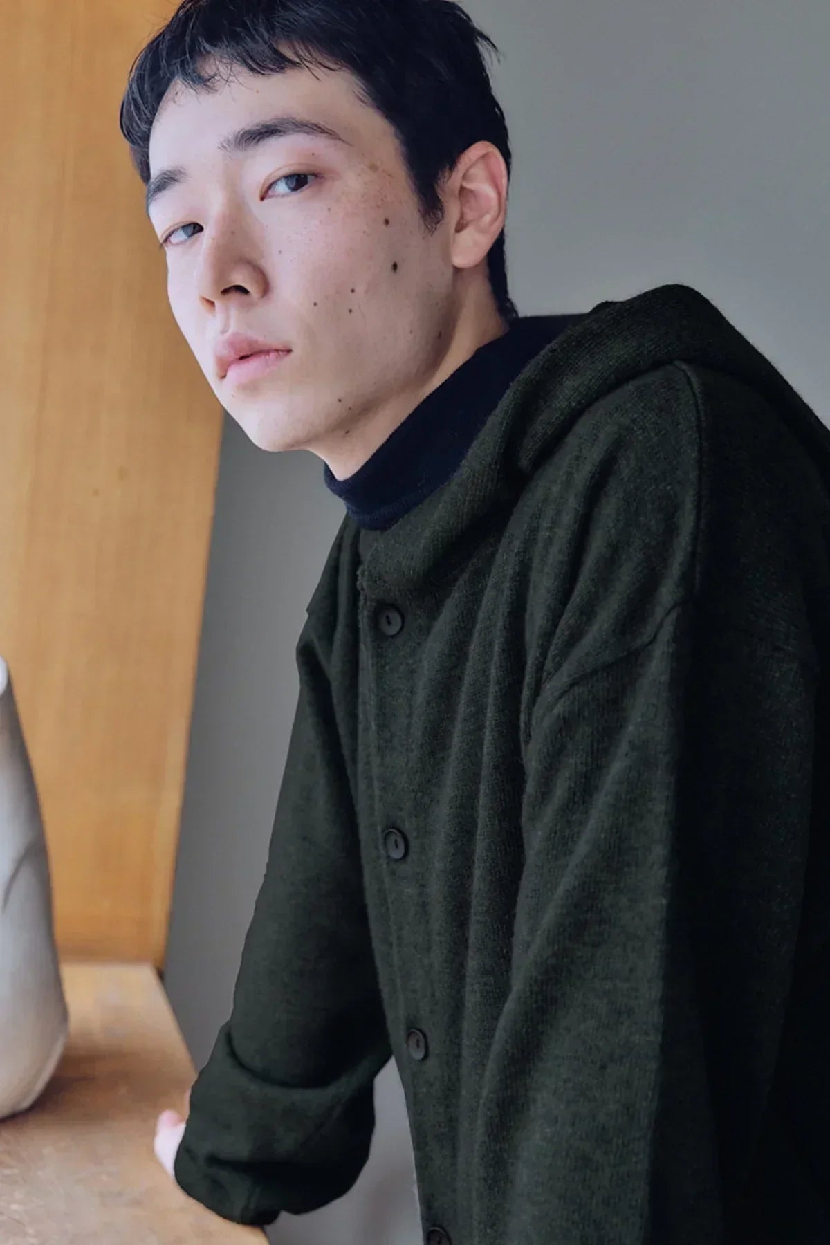 Close-up of a young man wearing a dark green knit hooded cardigan with black buttons indoors