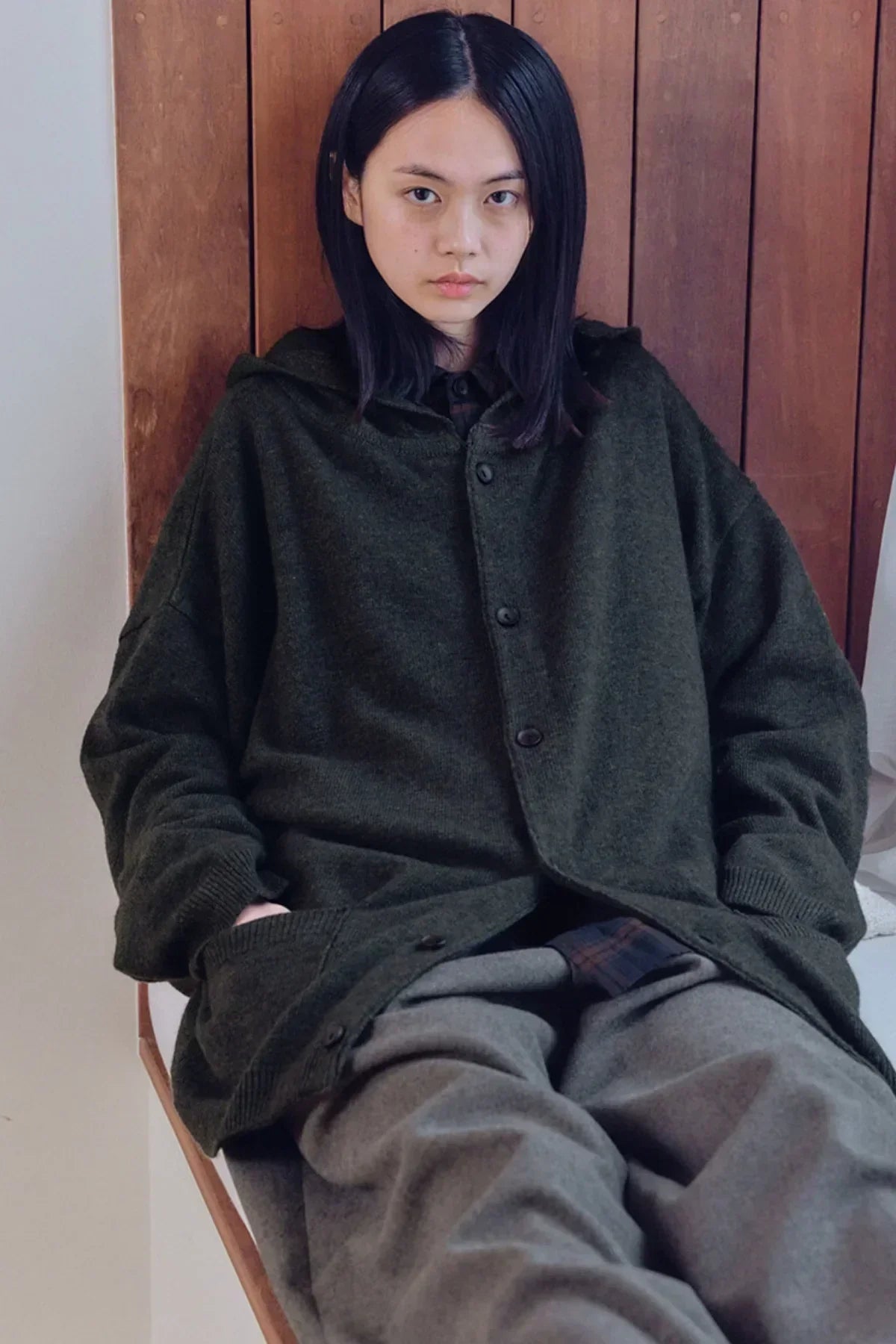 Young woman wearing a dark green unisex knit hooded cardigan sitting against wood panel backdrop