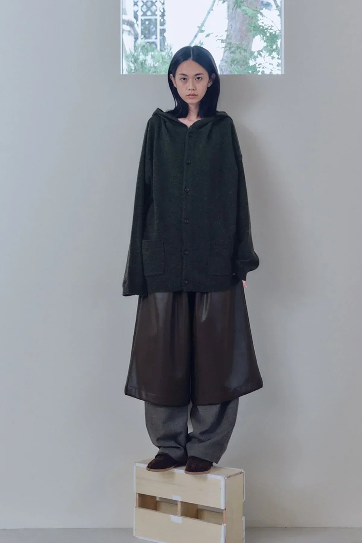 Model wearing oversized dark knit hooded cardigan and wide leather shorts standing on wooden crate