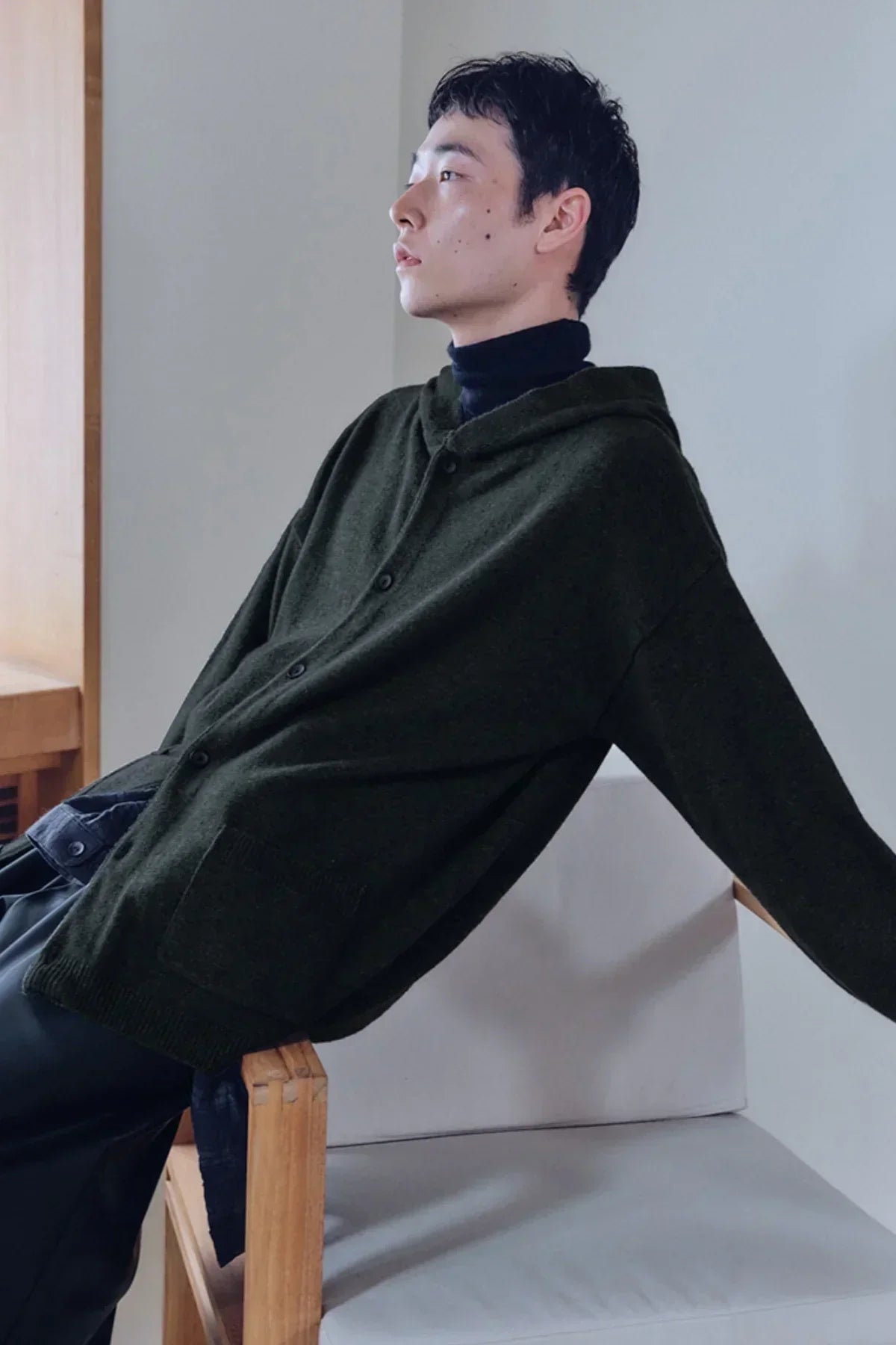 Man in khaki knit hooded cardigan sitting on a wooden chair in a minimal indoor setting