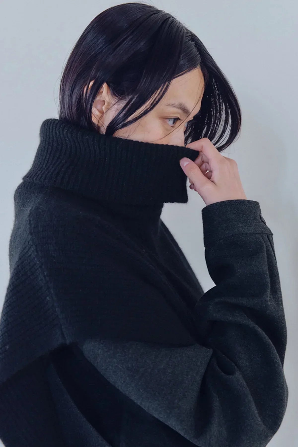 Close-up of person wearing black knit turtleneck vest and dark jacket against plain background