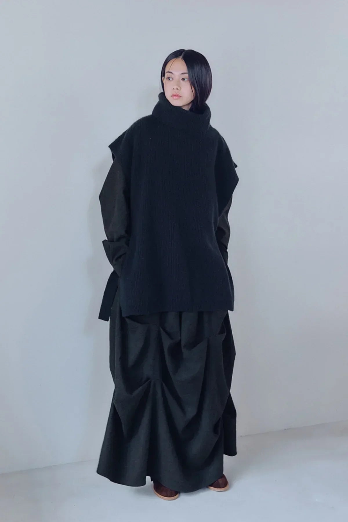 Model wearing black unisex knit turtleneck vest over dark oversized dress with draped skirt