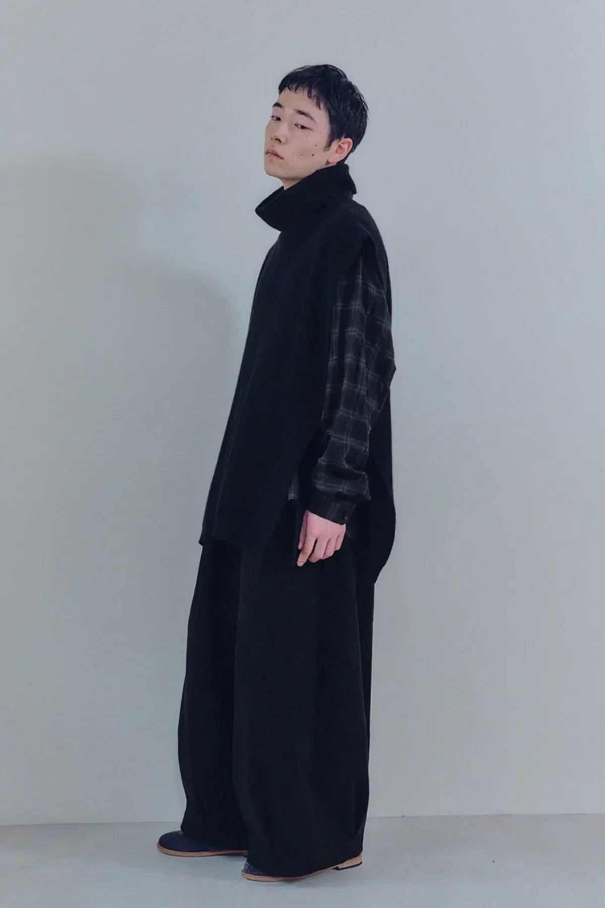 Unisex model wearing black knit turtleneck vest over plaid shirt and wide black pants in studio
