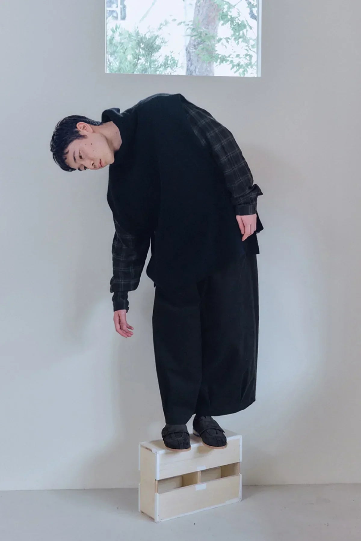 Person in black turtleneck knit vest over plaid shirt and wide black pants standing on wooden crate in white room