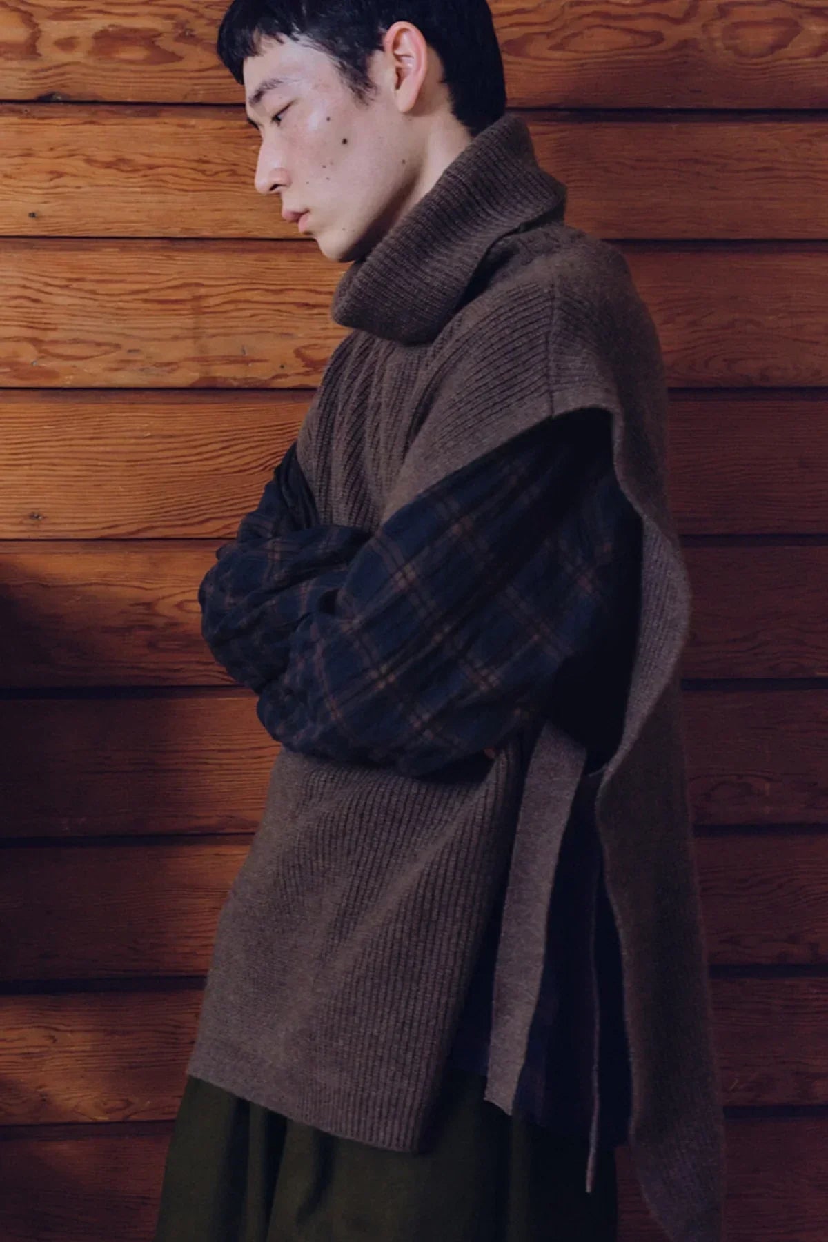 Side view of a person wearing a brown unisex knit turtleneck vest over a plaid shirt against a wooden wall
