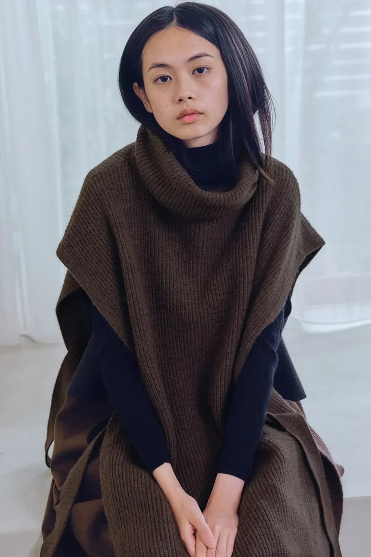 Woman wearing brown knit turtleneck vest layered over black long sleeves against white sheer curtain background