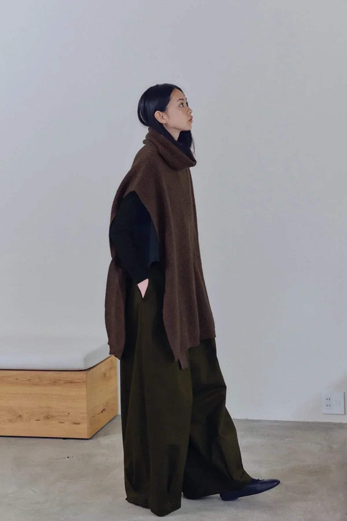 Woman wearing a brown knit turtleneck vest over black top and wide-leg olive pants, standing indoors