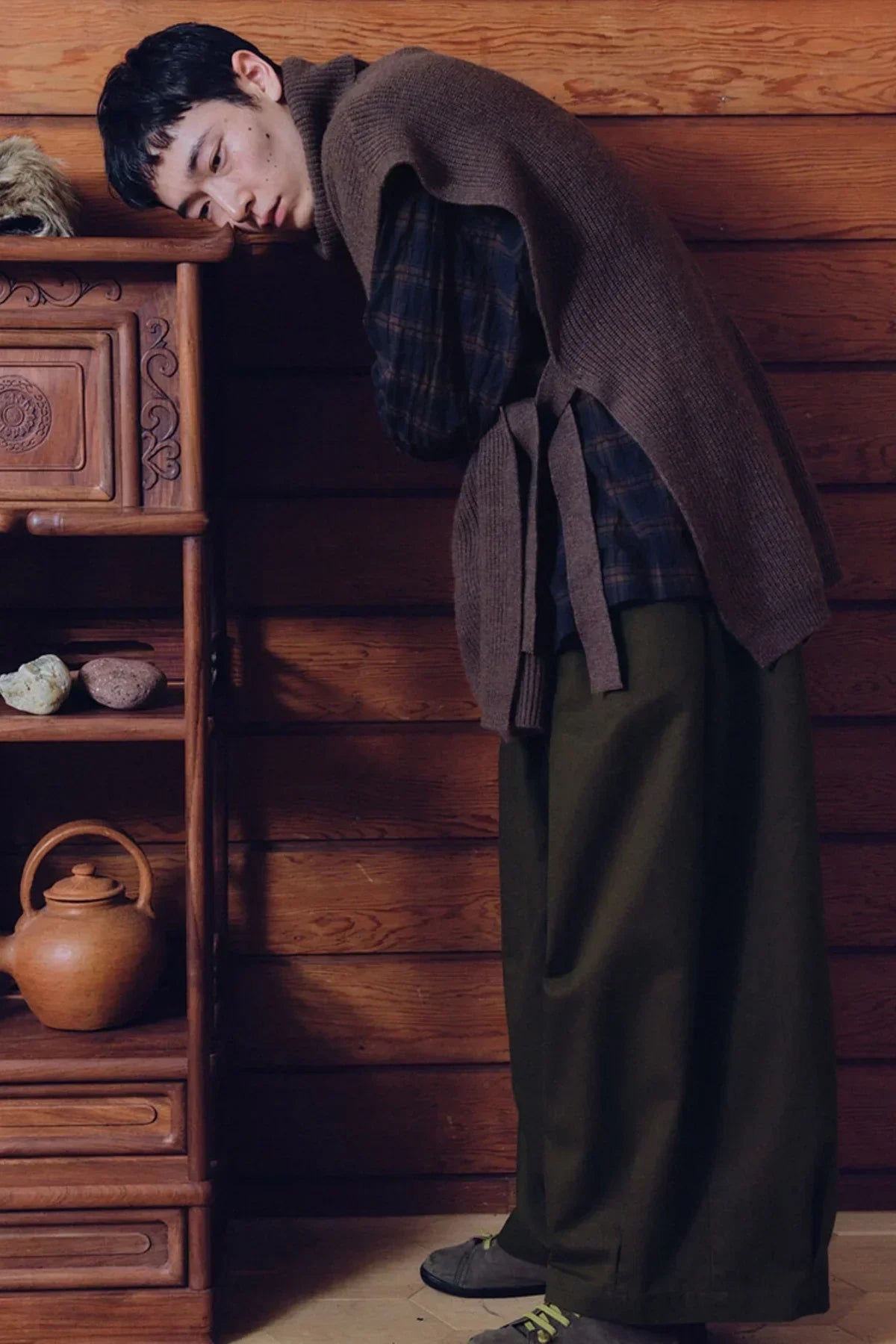 Man wearing brown knit turtleneck vest, plaid shirt, and loose olive pants leaning on wooden shelf in rustic room