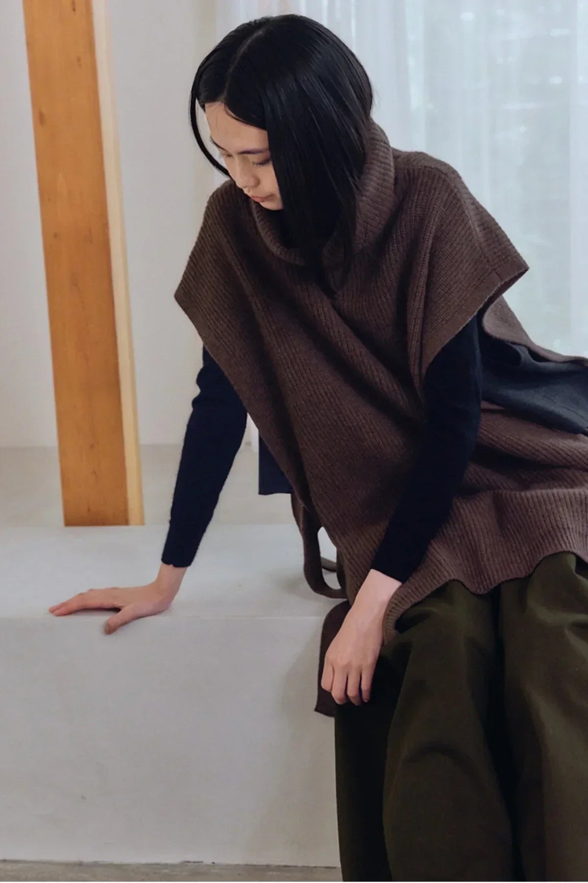 Person wearing brown unisex knit turtleneck vest over black top and green pants sitting indoors