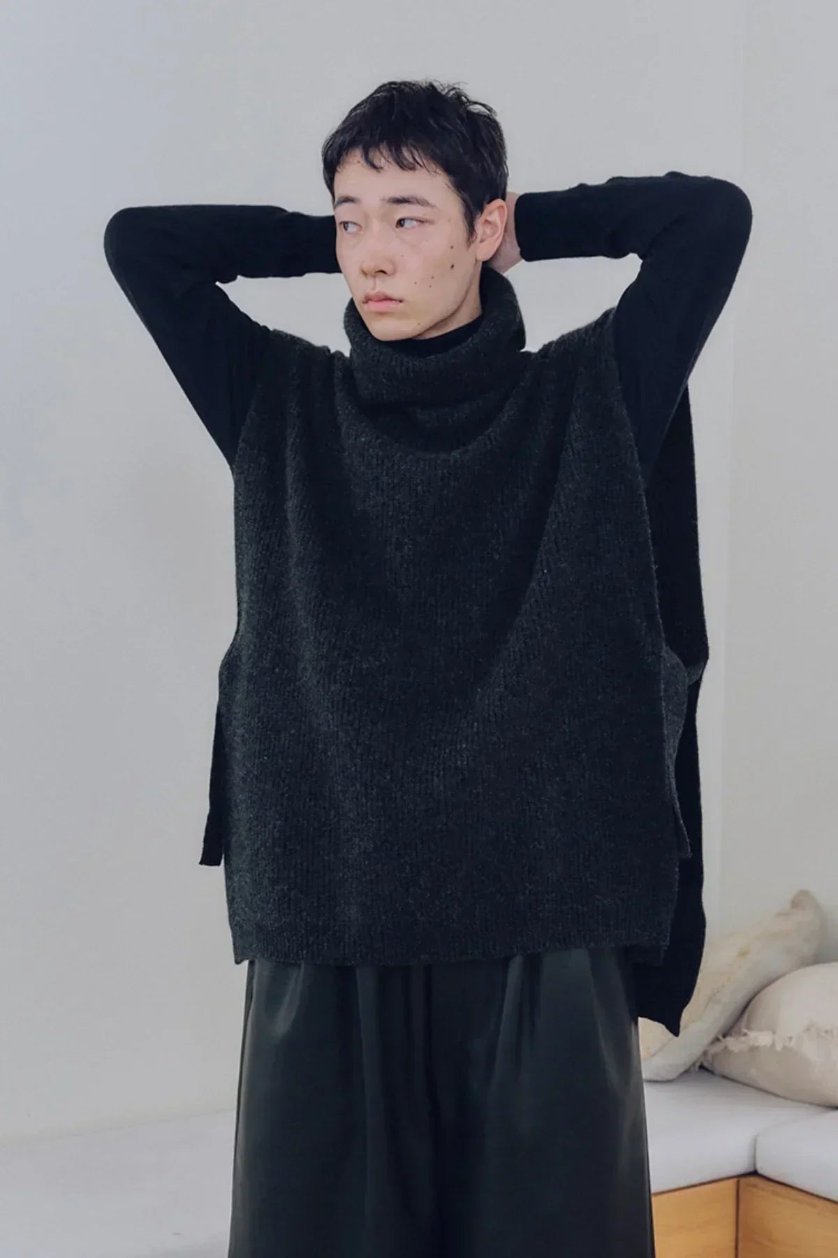Man wearing charcoal knit turtleneck vest over black long sleeves indoors with cushions in background
