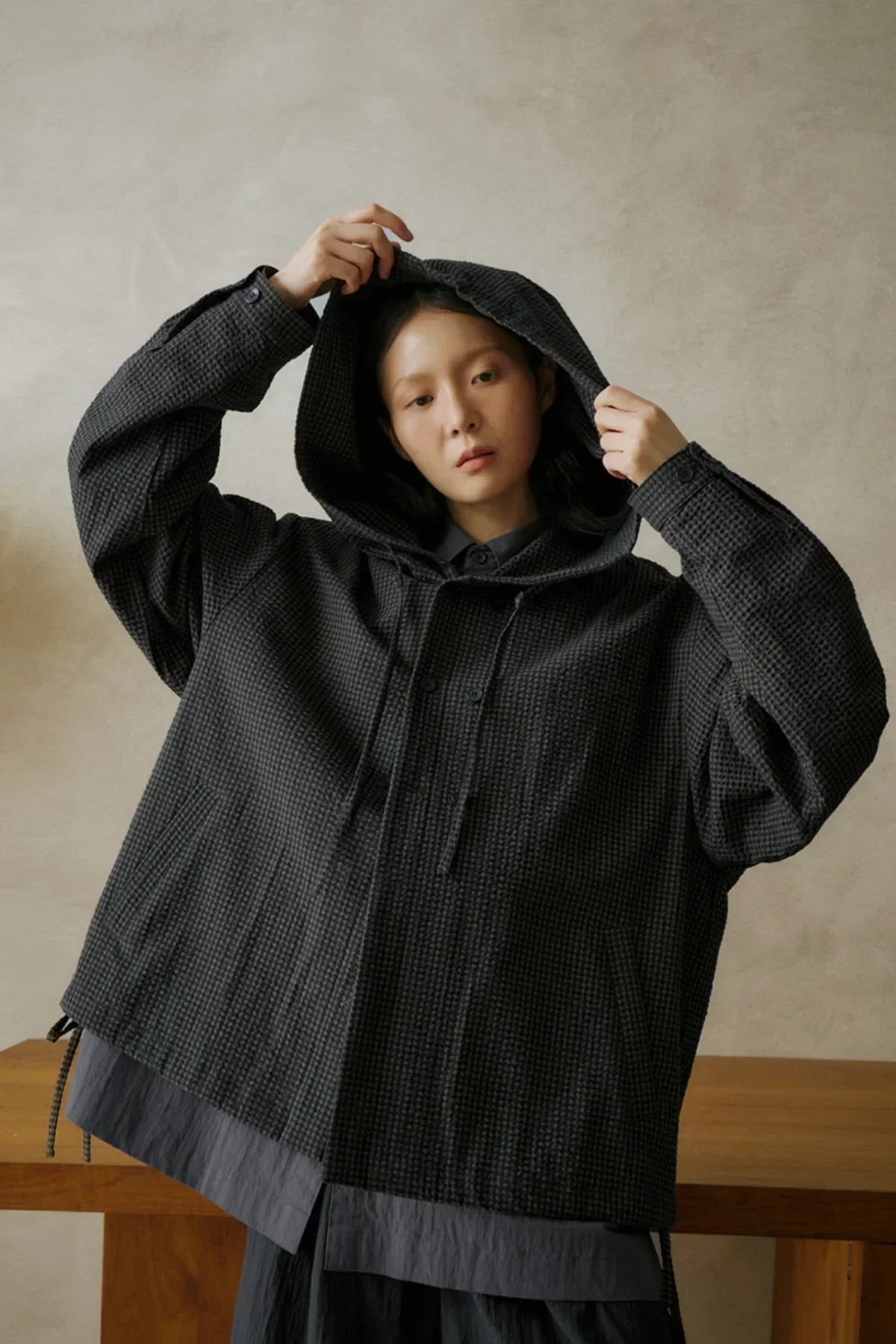 Woman wearing oversized black textured hooded jacket posing indoors against plain beige wall