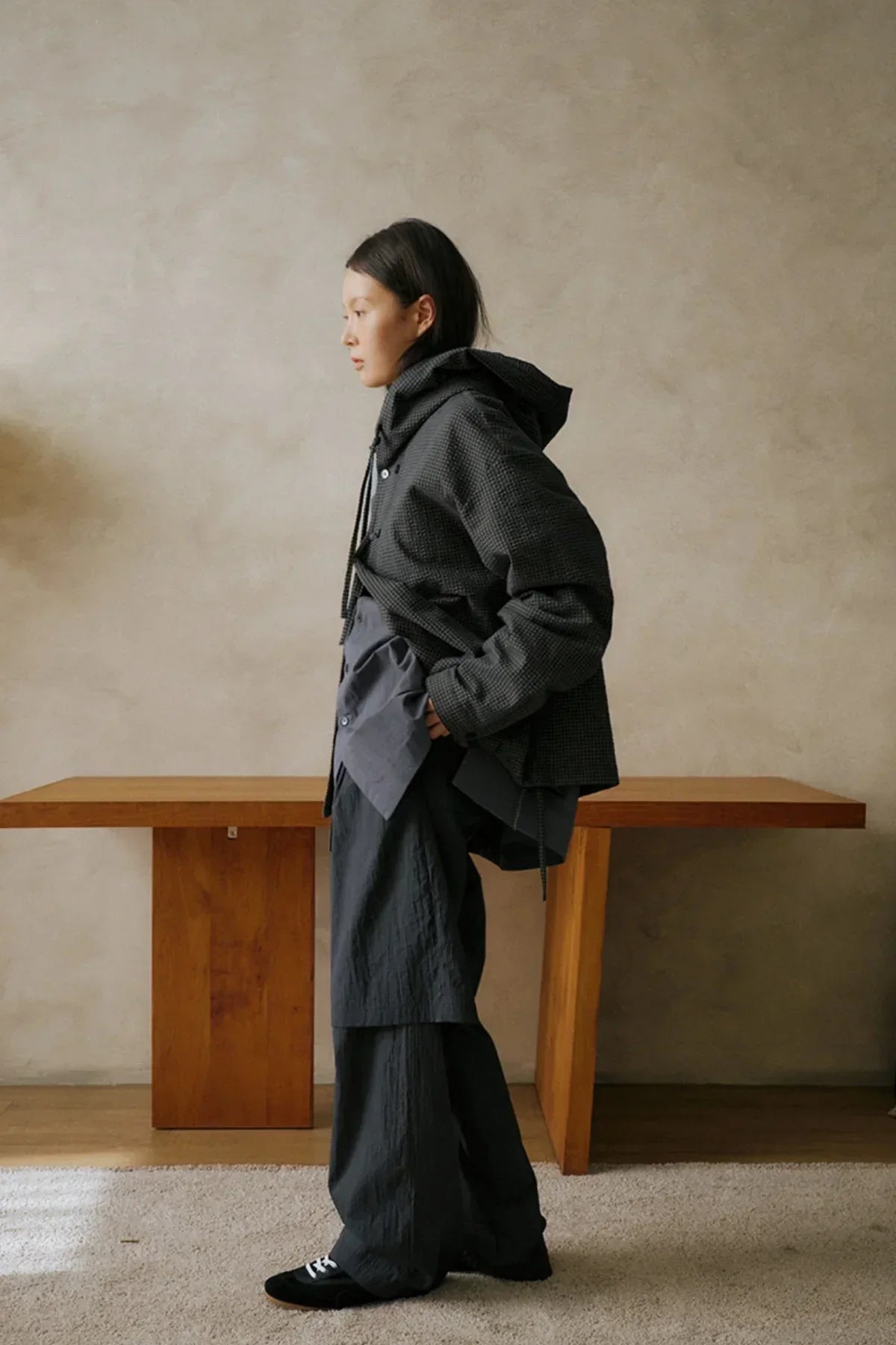 Side view of woman wearing black hooded jacket and wide pants standing indoors by wooden table