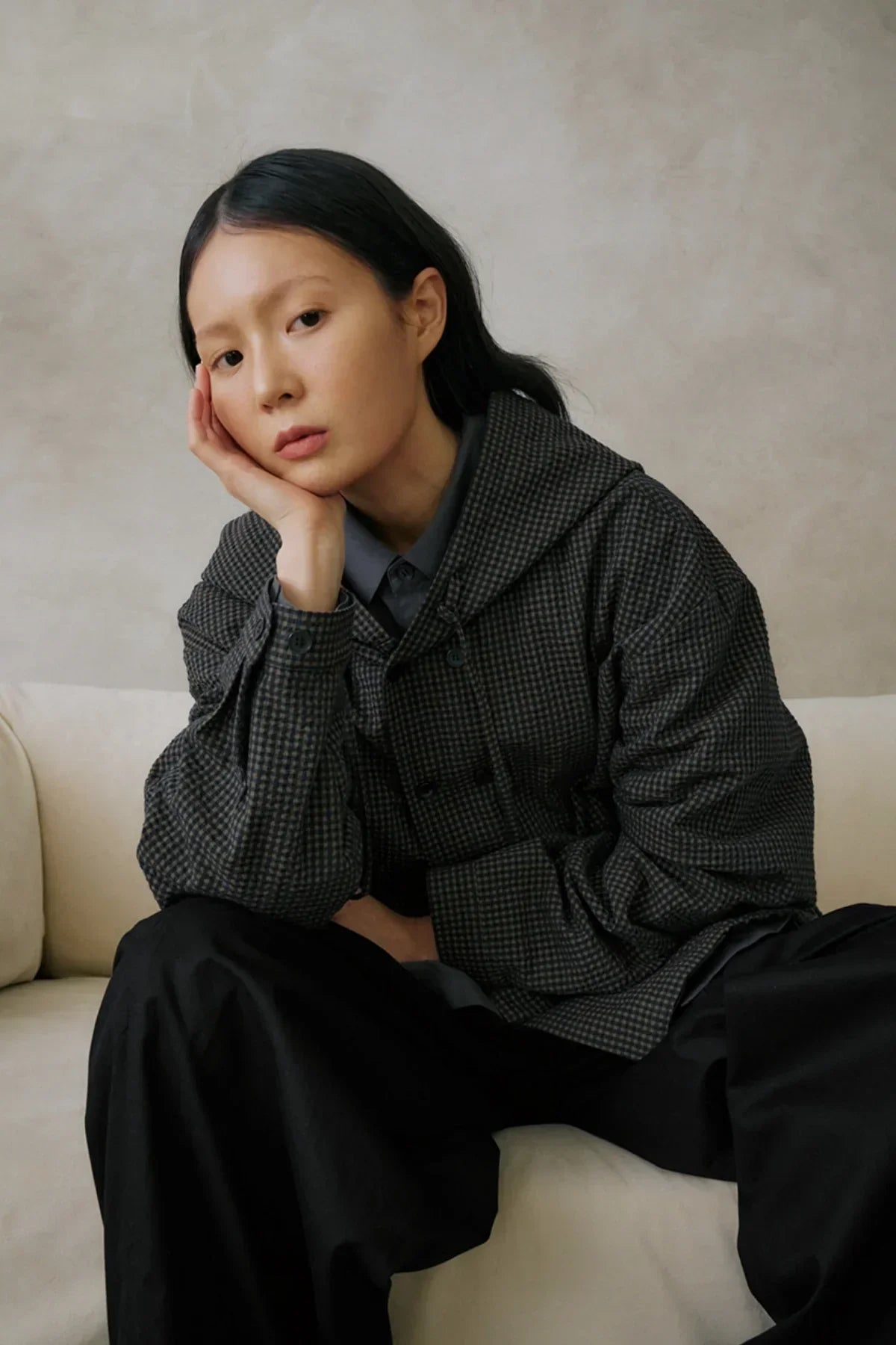 Woman seated on beige sofa wearing checkered hooded jacket and black pants against neutral wall