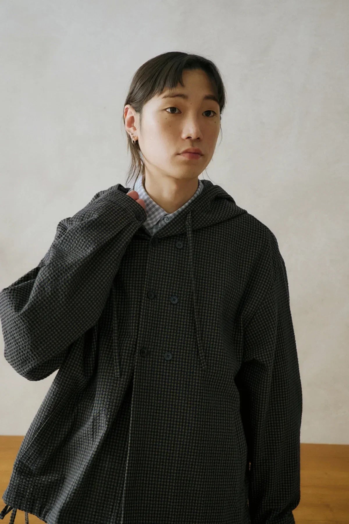 Young person wearing a dark checkered hooded jacket over a light blue checkered shirt in a neutral indoor setting