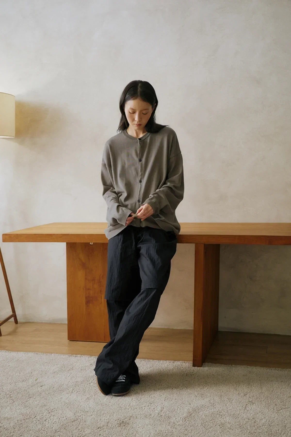 woman wearing loose black layered pants and oversized gray button-up cardigan leaning on wooden table in minimal room