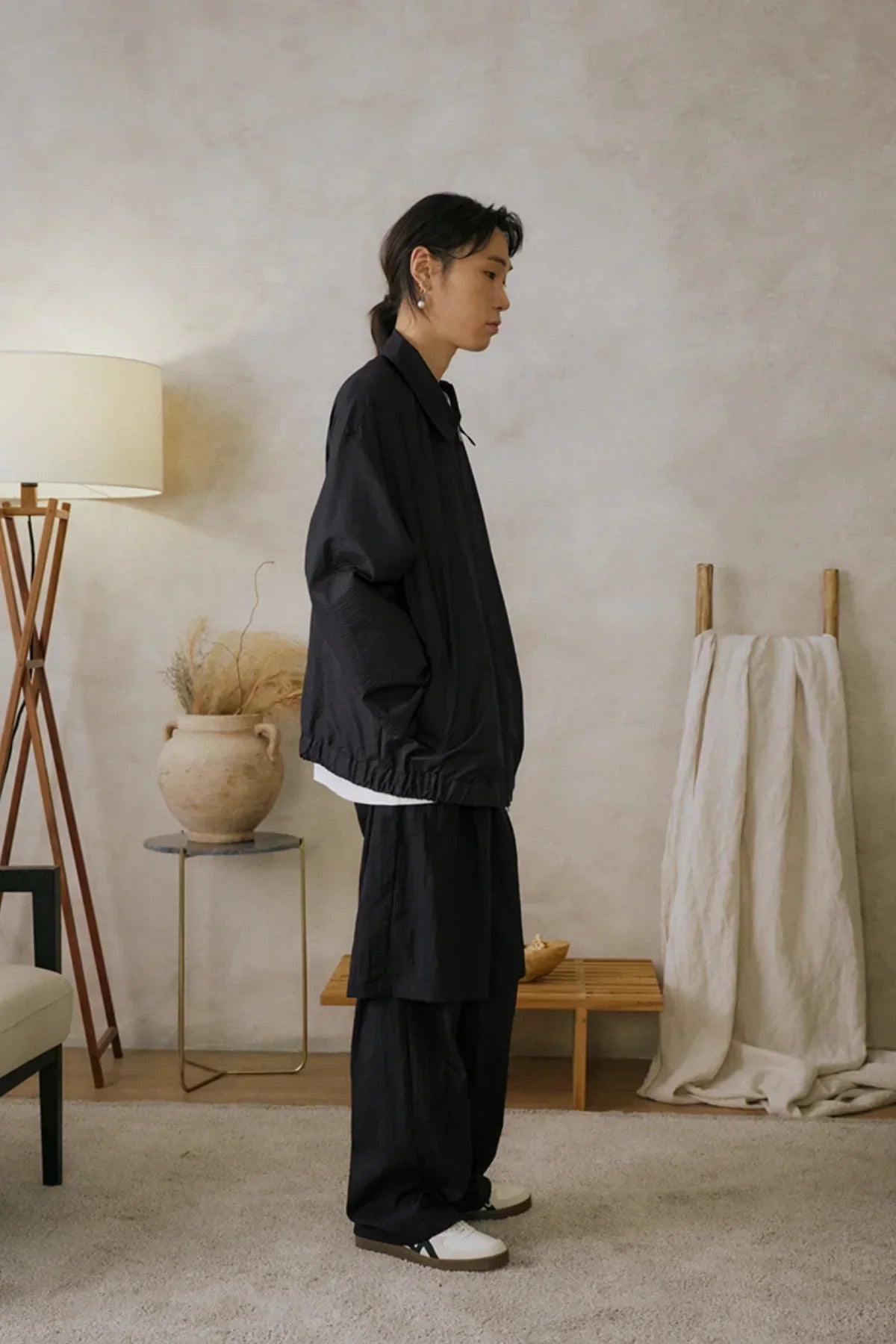 Profile of model wearing black loose layered pants and jacket in a minimalist room with beige walls