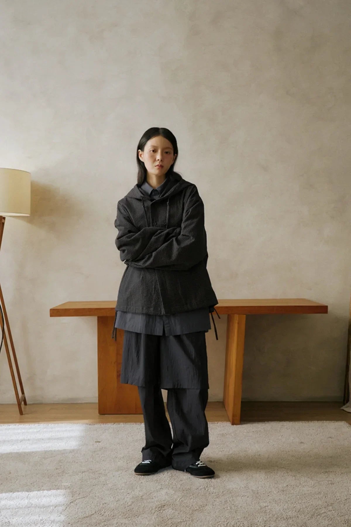 Person wearing dark layered unisex pants and jacket standing indoors by wooden table and lamp