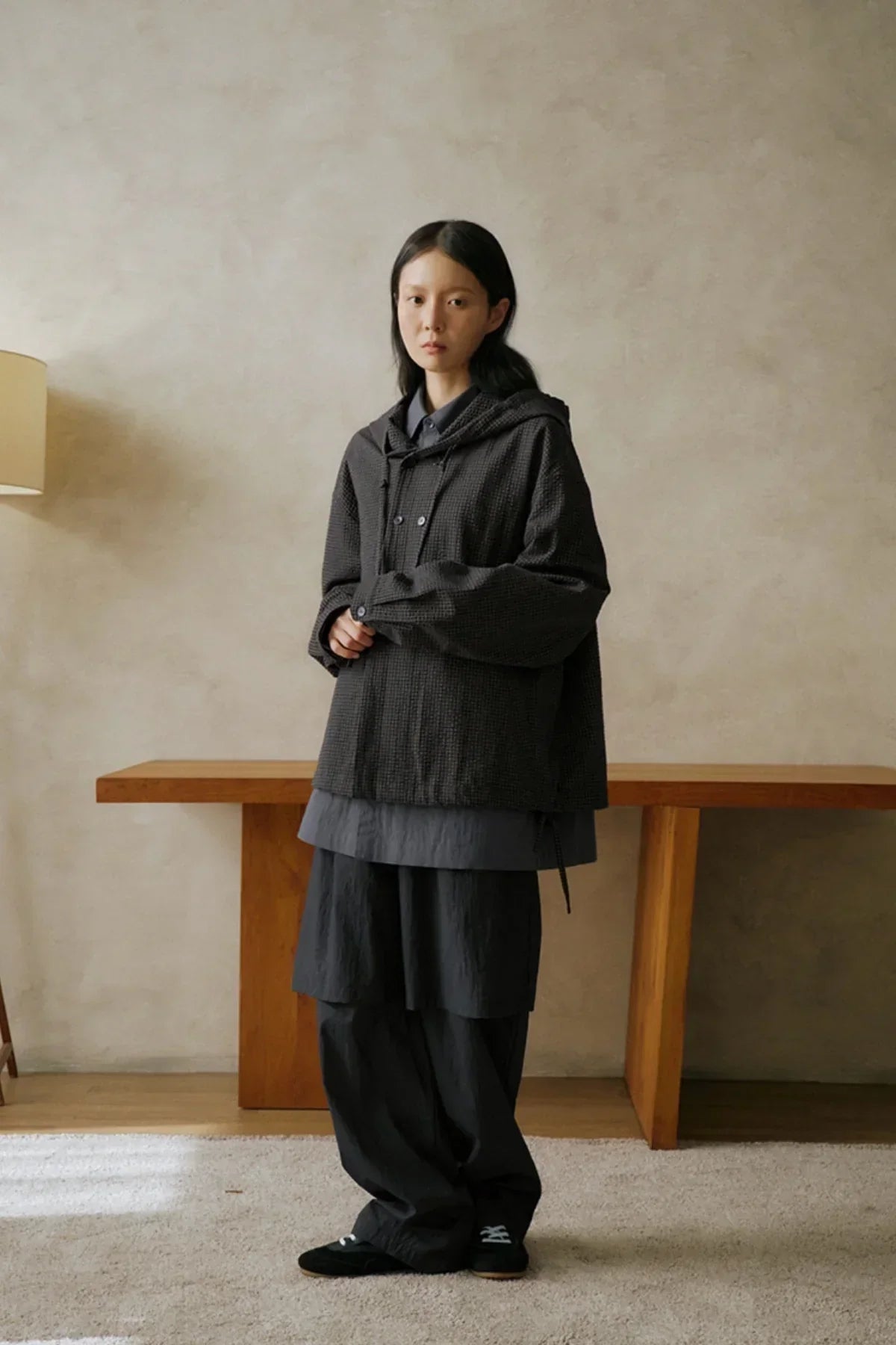 Woman wearing layered dark gray oversized hoodie and pants standing indoors by wooden table and beige wall