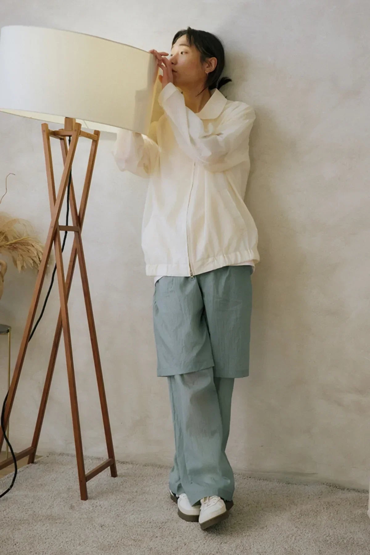 Person wearing unisex layered mint green pants and white jacket adjusting a floor lamp indoors