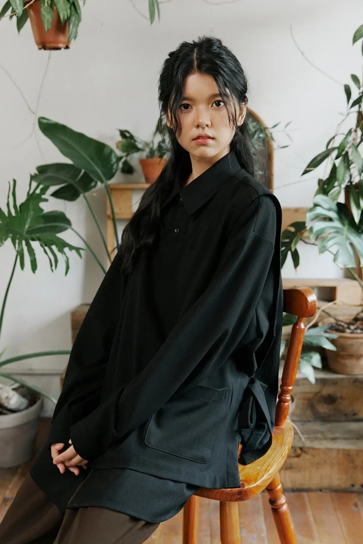 Young woman with long black hair wearing a loose black shirt sitting on a wooden chair indoors surrounded by green potted plants