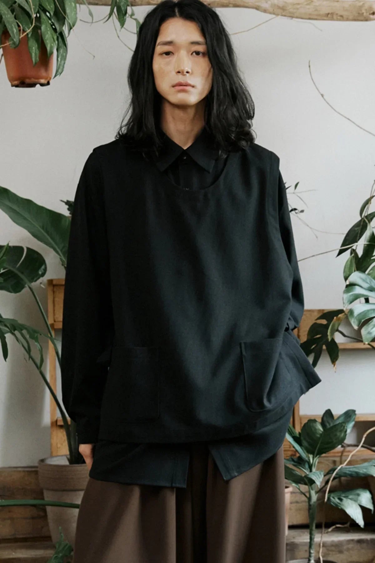 Young person with long hair wearing oversized black shirt and dark brown pants indoors with green plants