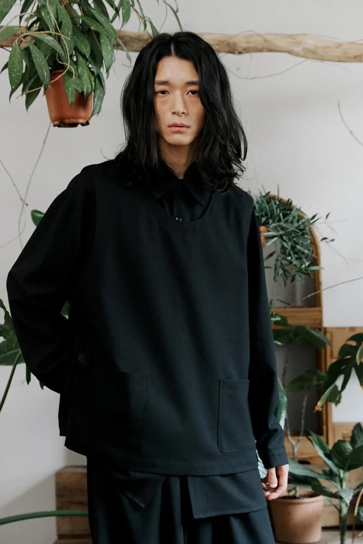 Unisex black layered shirt and vest outfit worn by model with long hair in indoor plant-filled setting