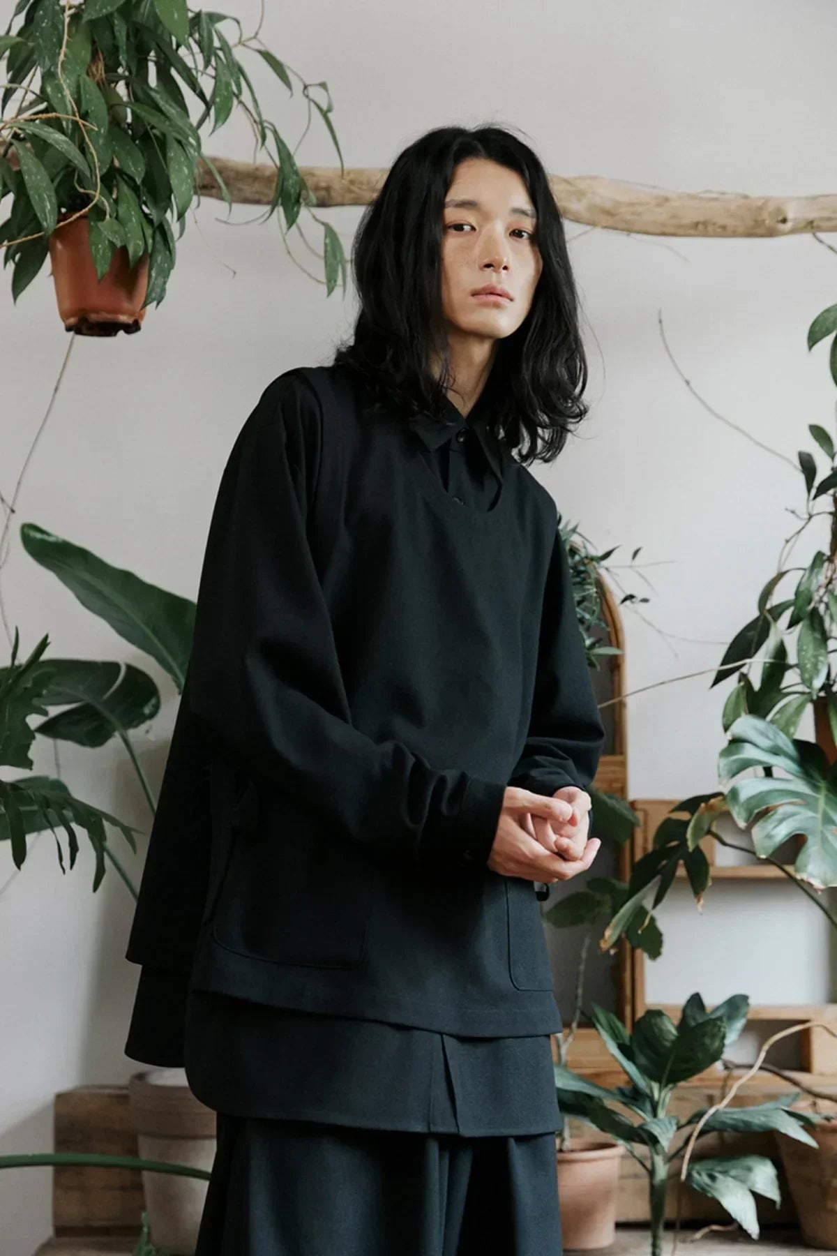 Young Asian model wearing layered black unisex shirt and vest surrounded by green indoor plants
