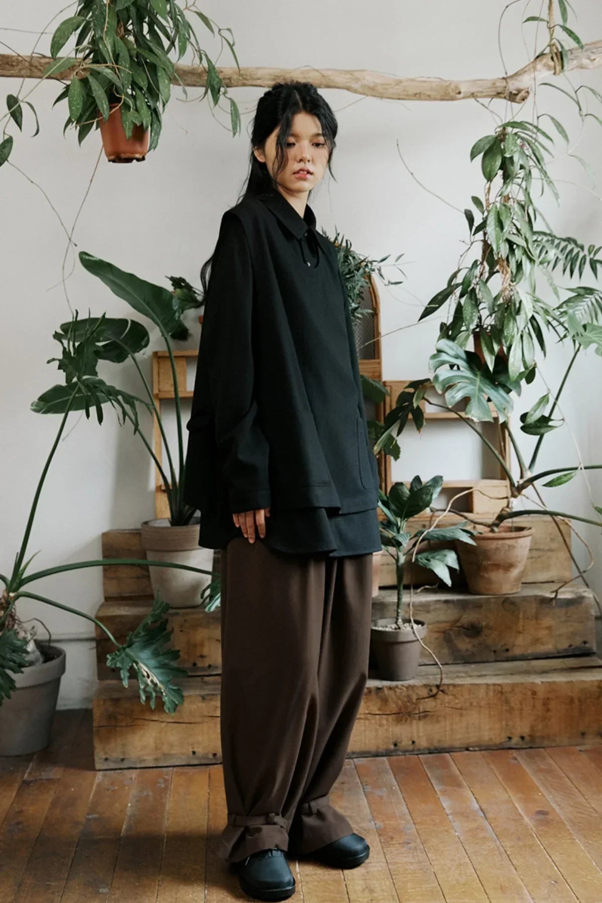 Model wearing oversized black shirt and loose brown pants standing indoors with potted green plants