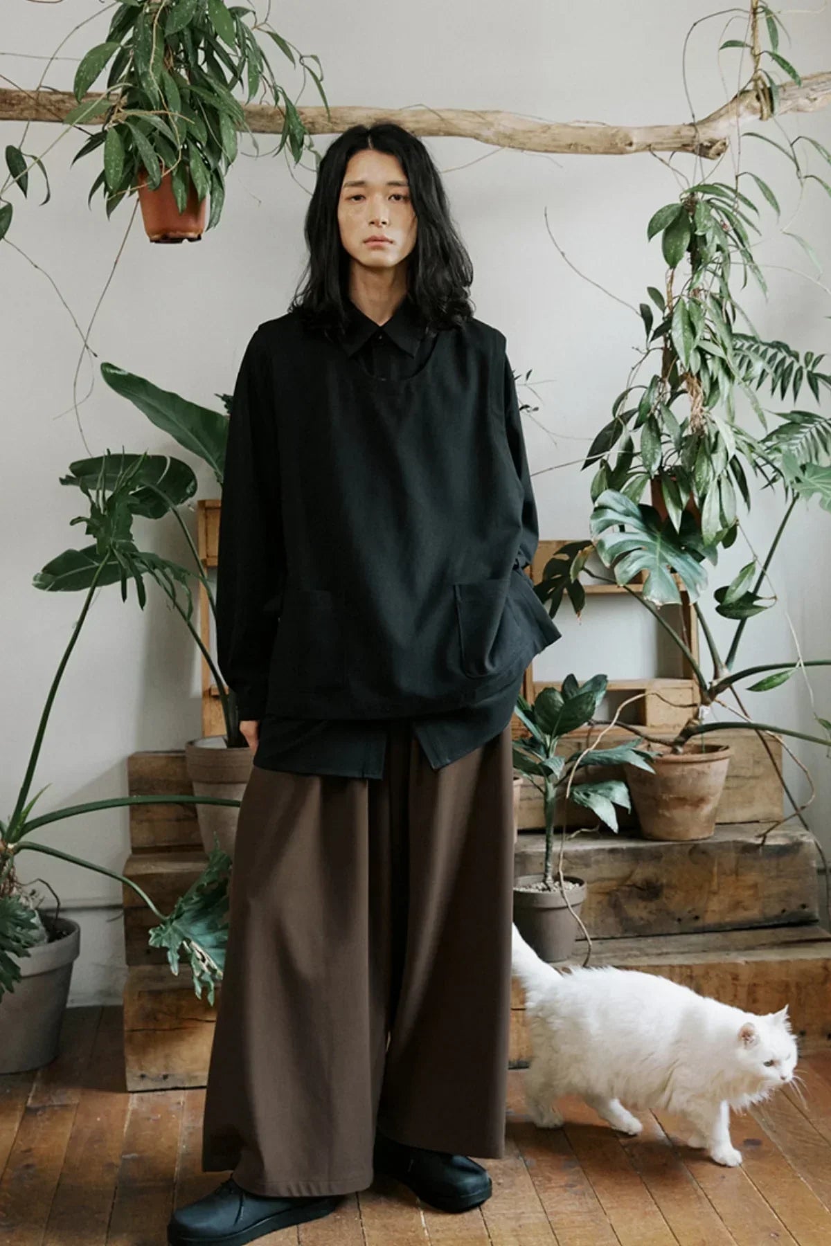 Unisex fashion model in black oversized shirt and brown wide pants standing indoors with houseplants and a white cat