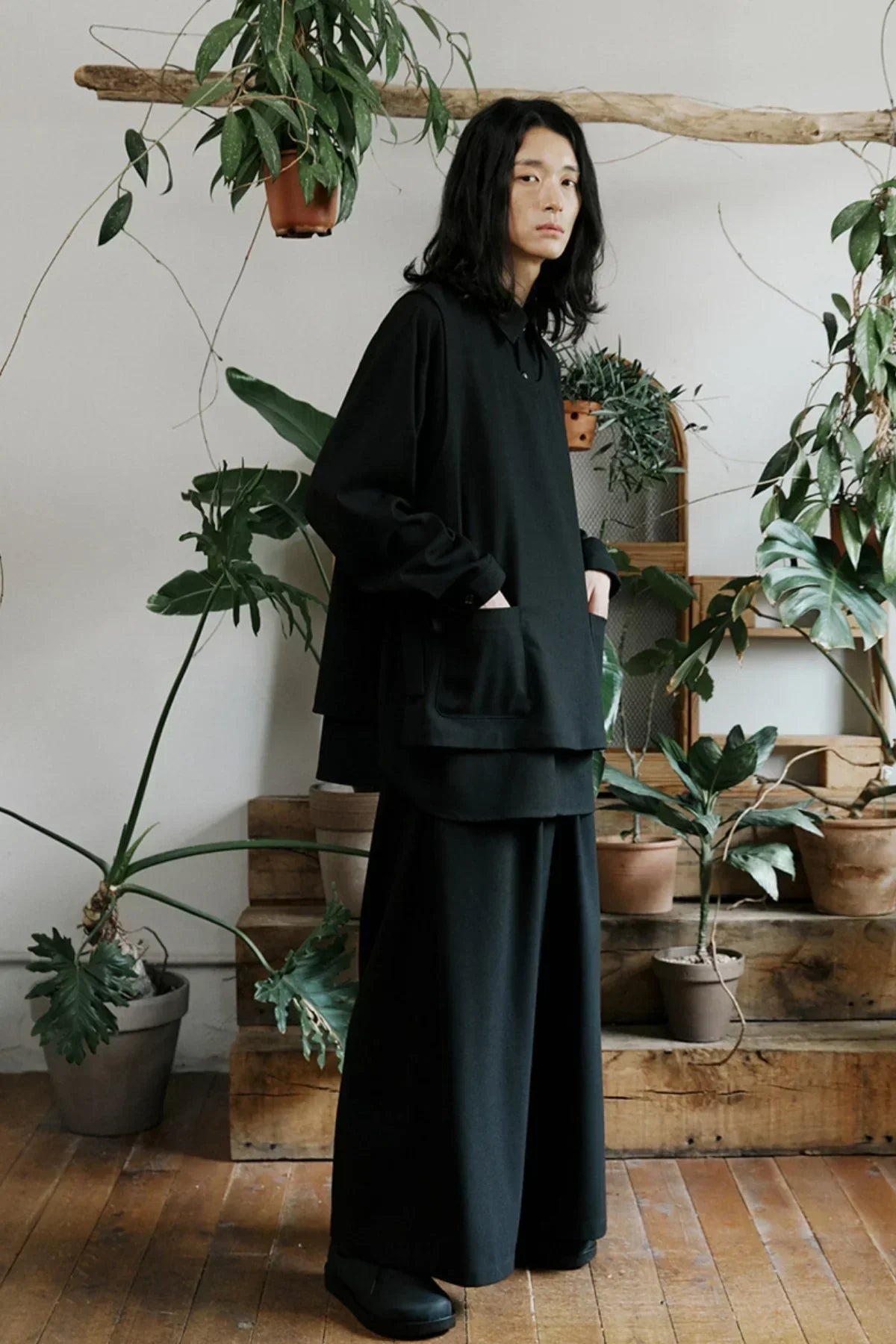 Model in black unisex long-sleeve shirt and wide-leg pants posing indoors with indoor plants