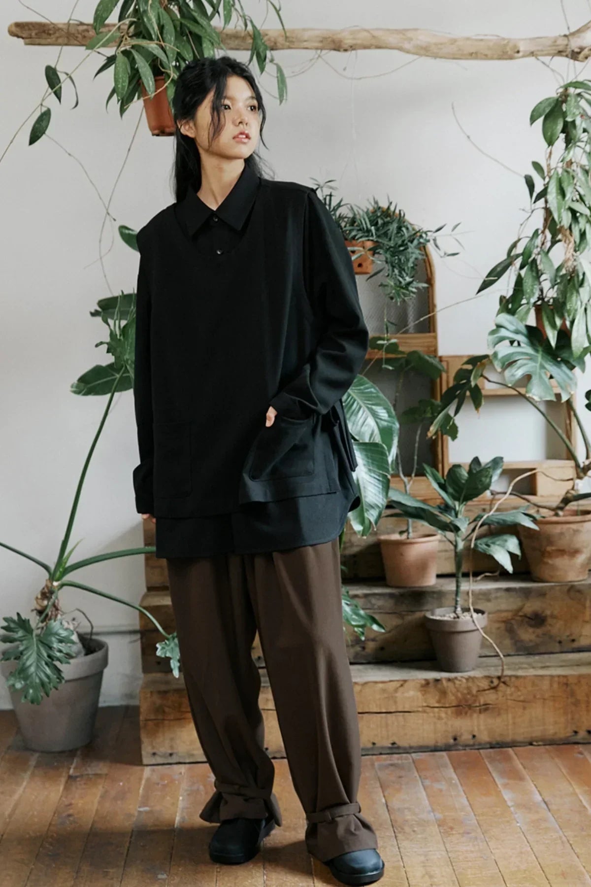 Woman wearing oversized black shirt and loose brown pants standing indoors among potted green plants
