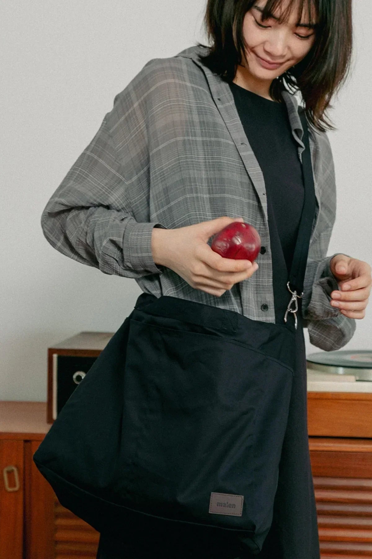 Woman holding red apple wearing plaid shirt and black dress with black unisex shoulder bag