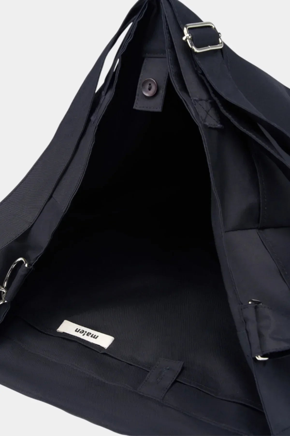Black unisex shoulder bag with adjustable strap, open flap showing interior label and button closure
