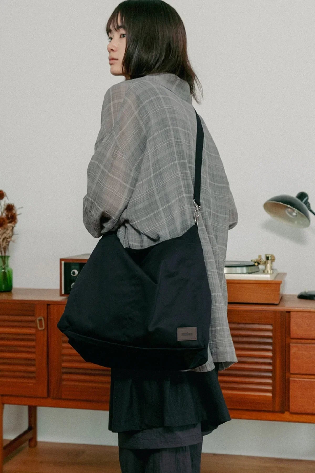 Woman wearing oversized plaid shirt and carrying a large black unisex shoulder bag indoors