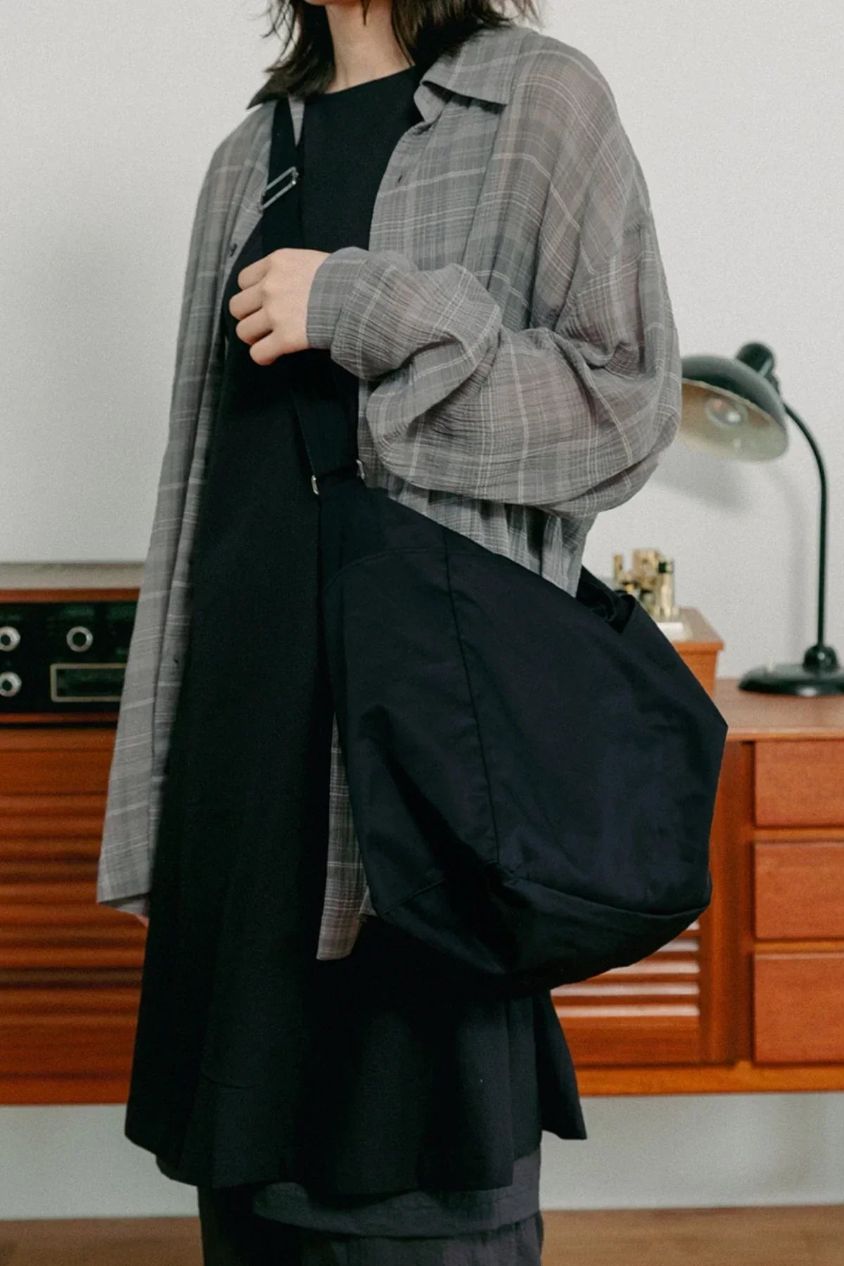 Person wearing black unisex shoulder bag with adjustable strap indoors, dressed in black and gray plaid shirt