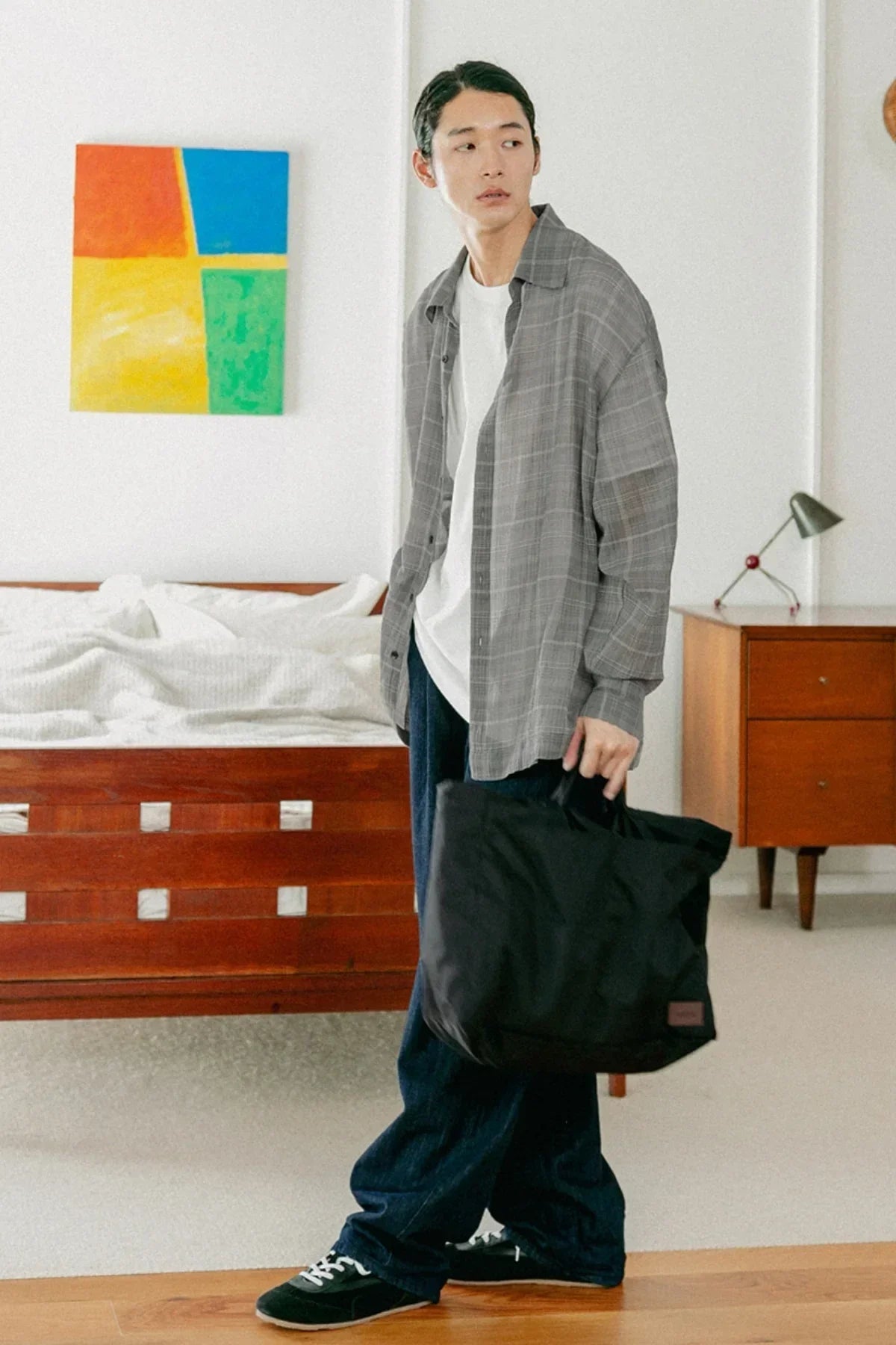 Young man in casual gray shirt and jeans holding black unisex shoulder bag in modern bedroom