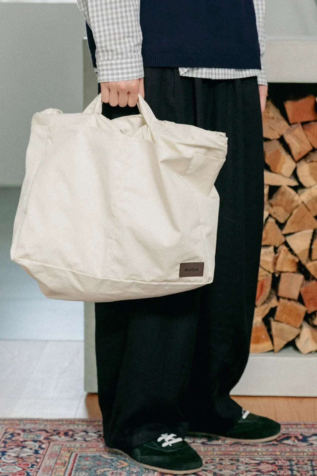 Person holding cream-colored unisex shoulder bag indoors by stacked firewood