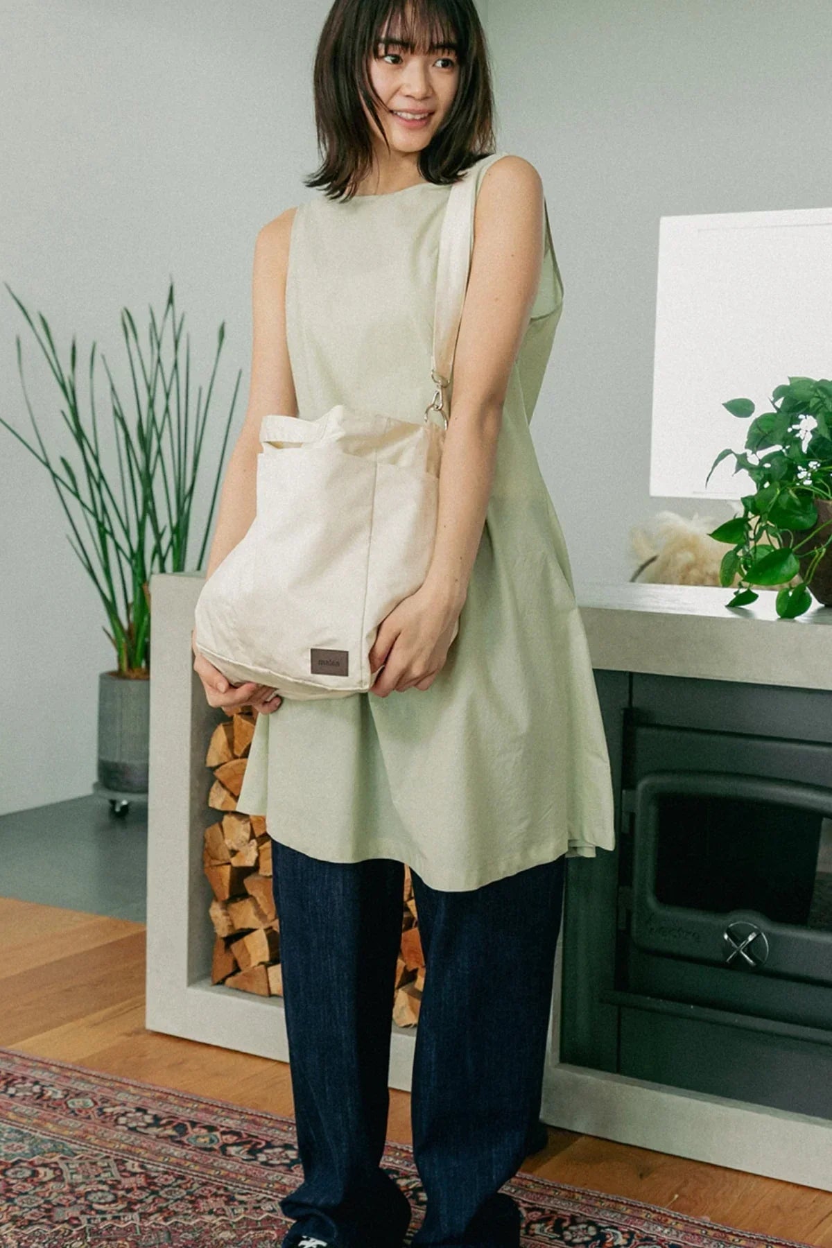 Woman wearing casual outfit with cream unisex shoulder bag standing indoors near fireplace and plants