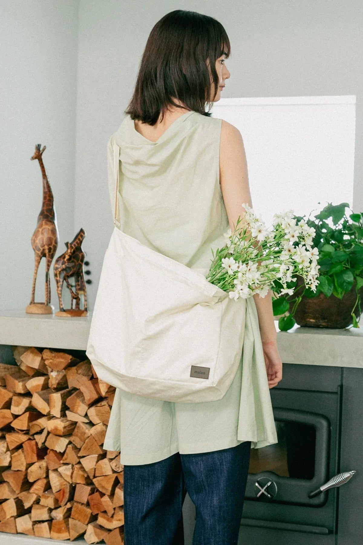Woman wearing unisex cream shoulder bag holding white flowers in modern cozy room with wood and plants