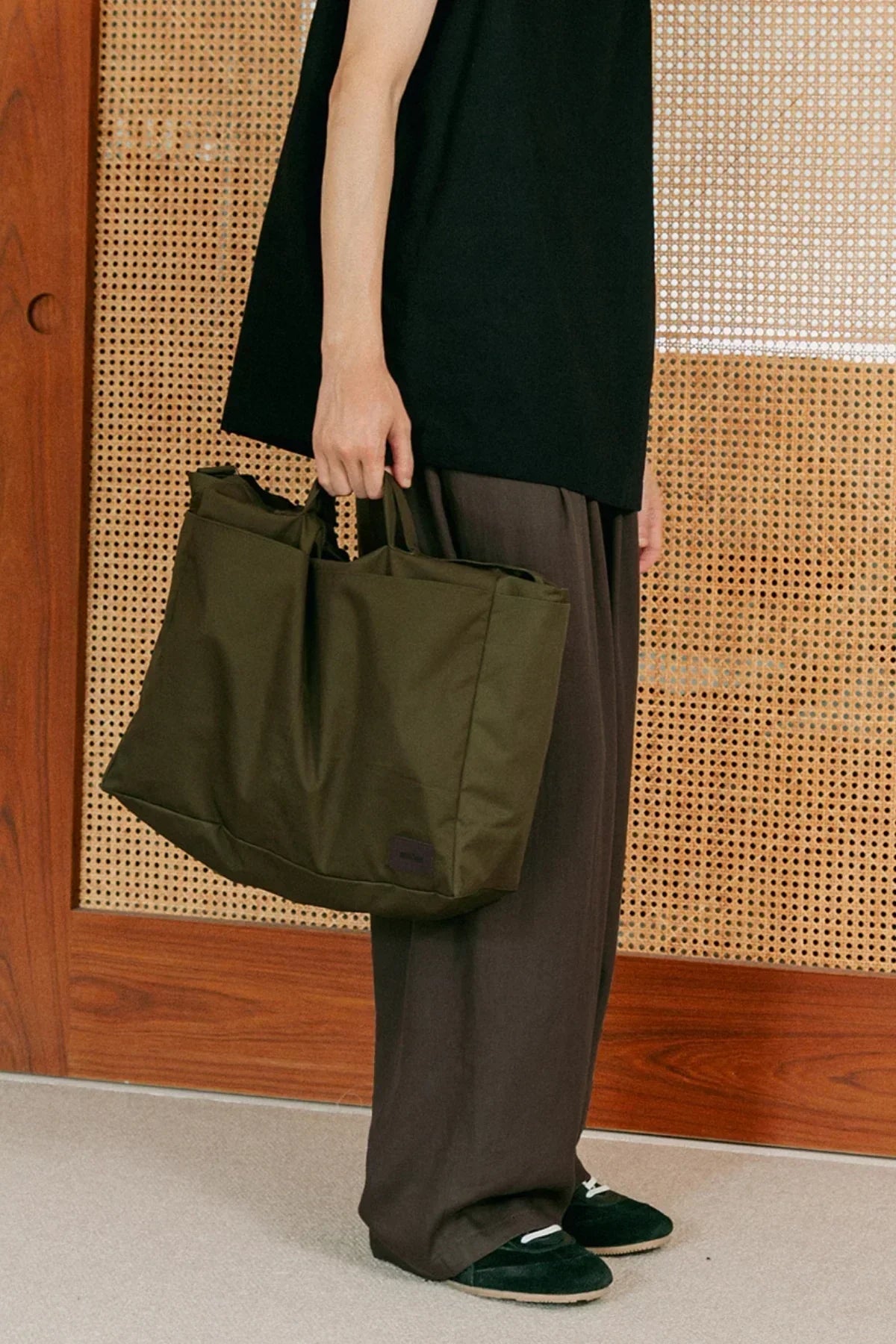 Person holding a khaki unisex shoulder bag wearing black top, brown pants, and black shoes indoors
