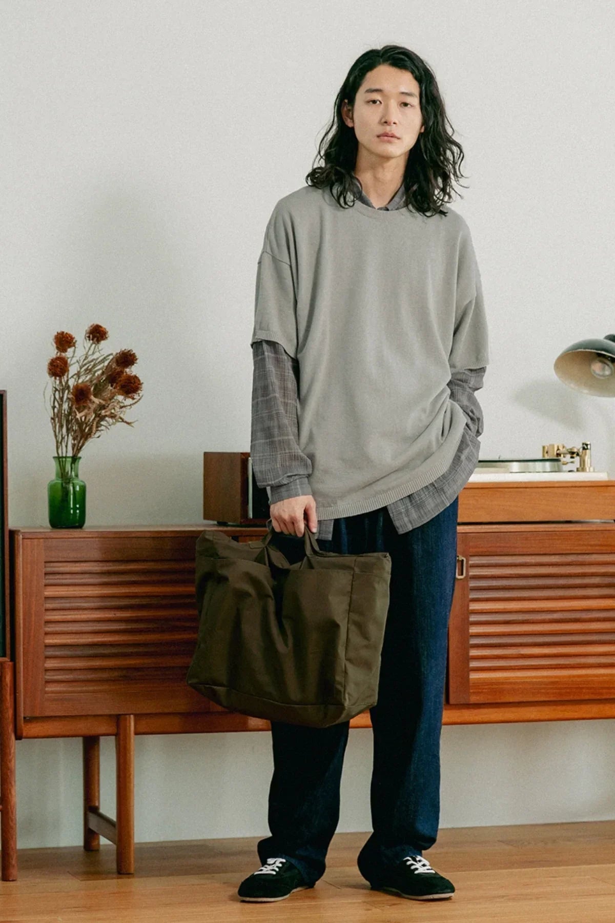 Man in casual layered outfit holding khaki unisex shoulder tote bag indoors by wooden furniture