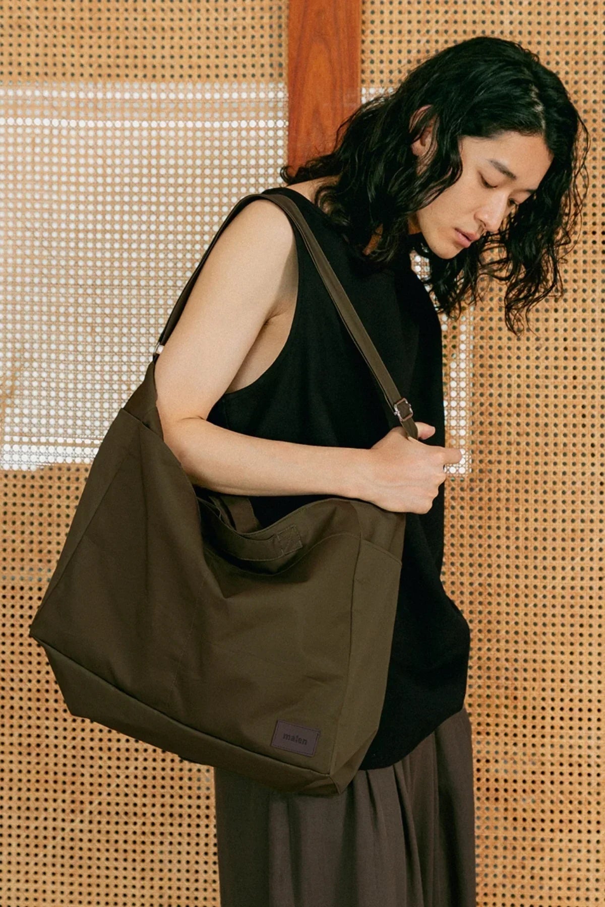 Young person with long hair wearing black sleeveless top carrying large khaki unisex shoulder bag against wooden patterned wall