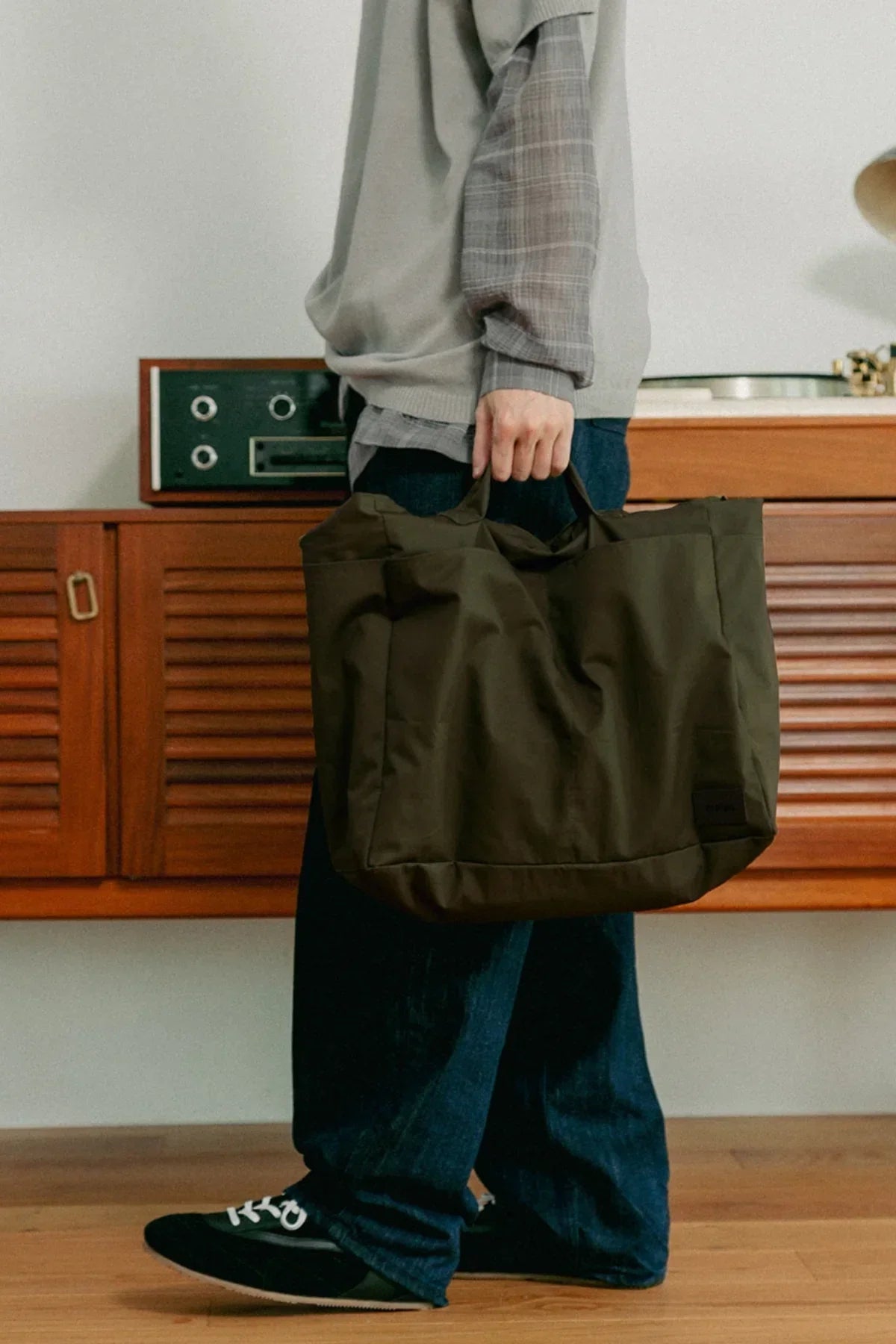 Person holding a khaki unisex shoulder bag indoors with casual outfit and wooden furniture background