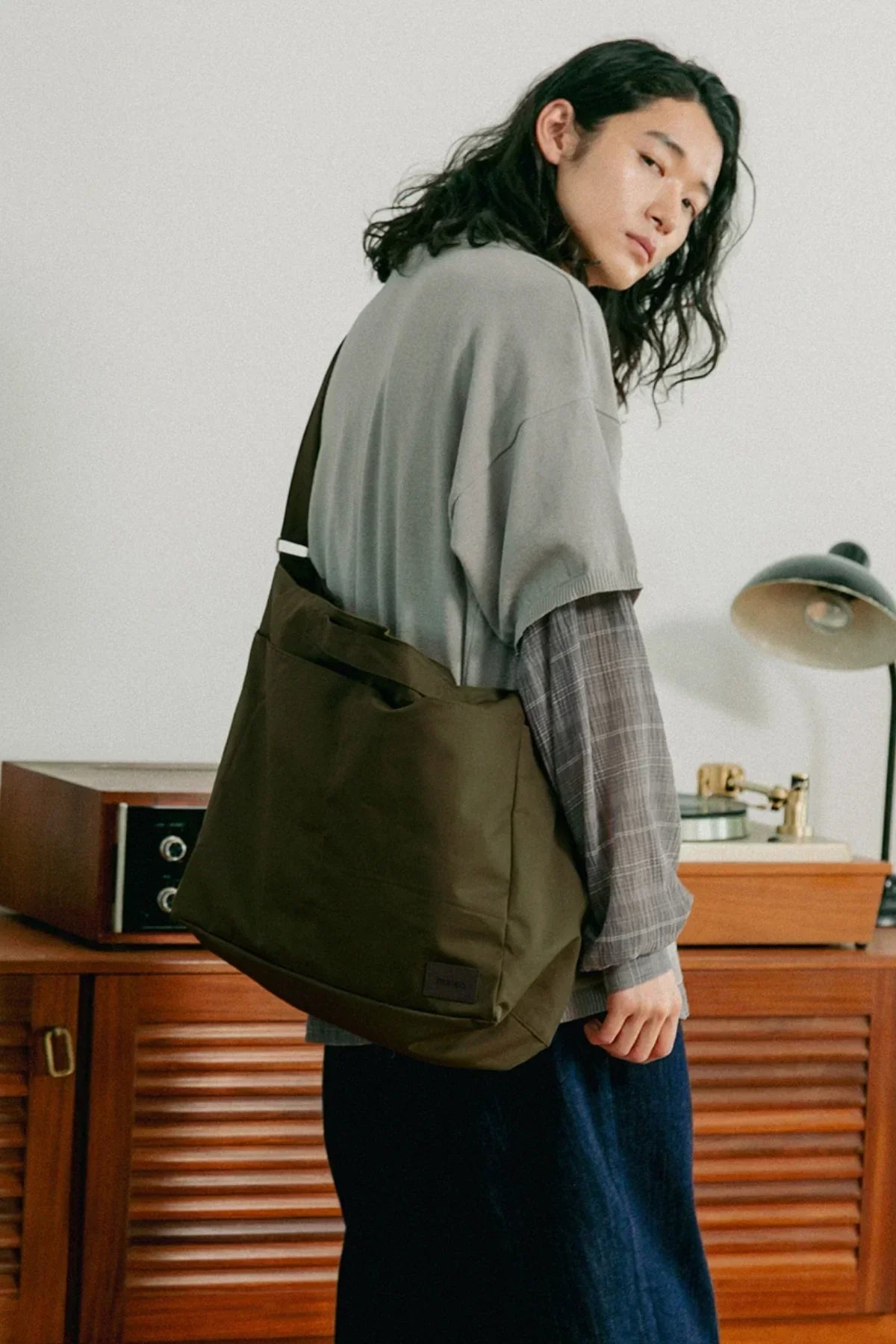 Person with long hair wearing a grey top and carrying a khaki unisex shoulder bag indoors
